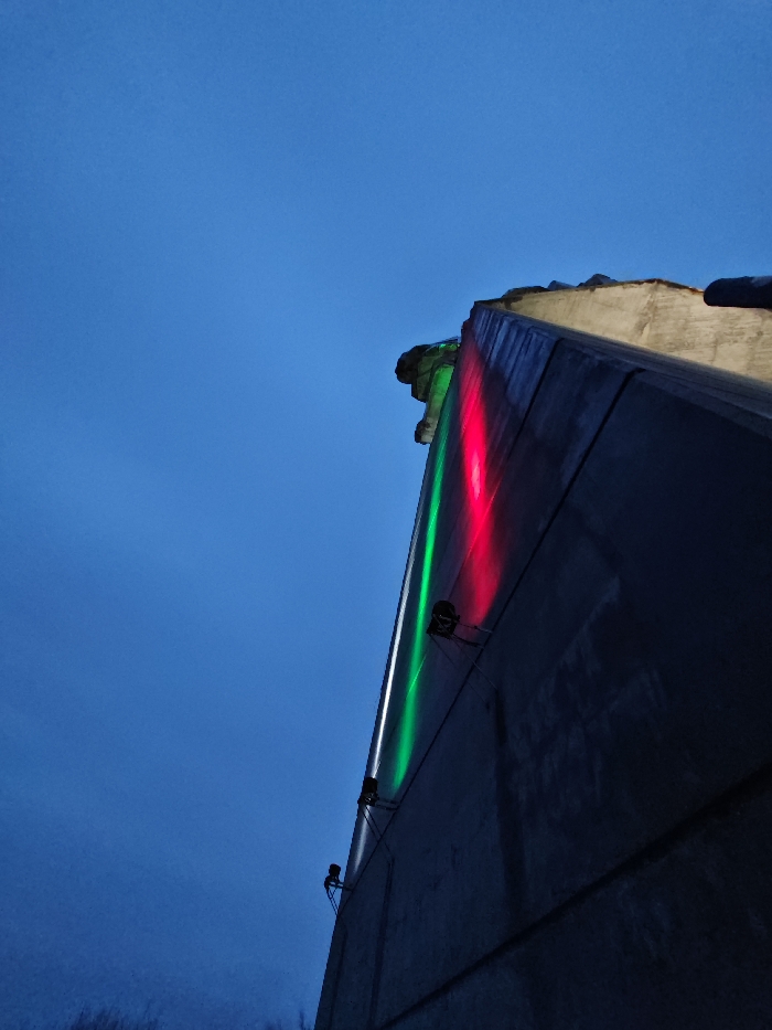 The image presents a low-angle, upward-looking view of a large concrete structure, identified as a section of the Monument to 1300 Years of Bulgaria in Shumen, Bulgaria, observed at twilight or night. The prominent feature is a tall, dark gray concrete wall, illuminated vertically by three distinct bands of artificial light: green on the left, white in the middle, and red on the right. Multiple small, dark spotlights are affixed to the concrete surface, projecting these colored beams upwards. The top edge of the concrete structure displays a rough, irregular profile and is illuminated by a subtle yellowish-white light from an unseen source. The upper and left portions of the frame consist of a clear, deep blue sky. Indistinct, dark shapes consistent with out-of-focus vegetation are marginally visible in the lower-left corner of the image. No individuals, animals, or vehicles are present.