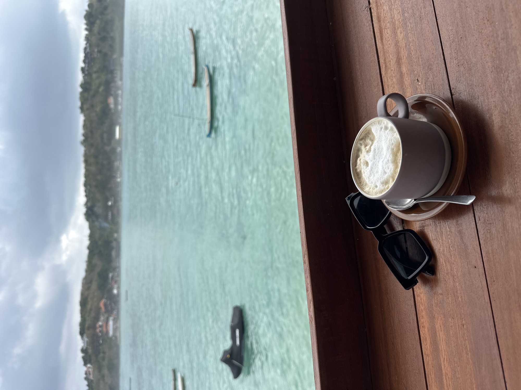A cup of coffee with frothy milk and a spoon rests on a saucer on a wooden table. To the left of the coffee, a pair of black sunglasses is placed. The table is in the foreground, with a blurred background showing the turquoise water of the ocean. Several small boats are visible on the water, and a lush green hill with some buildings is in the distance. The sky is overcast with gray clouds. This scene appears to be a view from a cafe or restaurant overlooking the water in Nusa Penida, Indonesia.