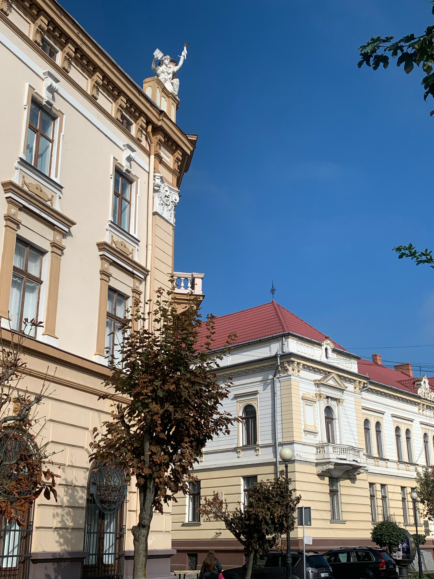 The image displays the historic architecture of Kolomyia, Ukraine, under a clear blue sky. The foreground features a building with a light yellow facade, adorned with intricate decorative elements and tall windows. A statue of a figure holding what appears to be a flag and a torch stands atop the building's corner, silhouetted against the sky. Adjacent to it, another building with a red-tiled roof and a similar architectural style is visible. Trees with autumn foliage are scattered across the scene, some with bare branches. In the lower portion of the image, the street level reveals parked cars and hints of pedestrian activity, with a person's back visible in the lower left corner, and another in the lower right. The overall impression is of a well-preserved historical district bathed in sunlight.