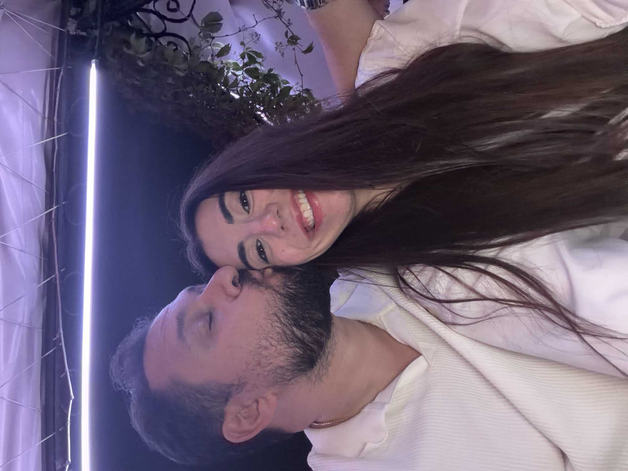A man and woman are posing closely together for a picture. The woman is smiling, while the man has his eyes closed and head tilted towards her. Both are wearing white shirts. The background includes a dimly lit area with some plants. The image does not provide enough information to determine a specific location in Sofia, Bulgaria.