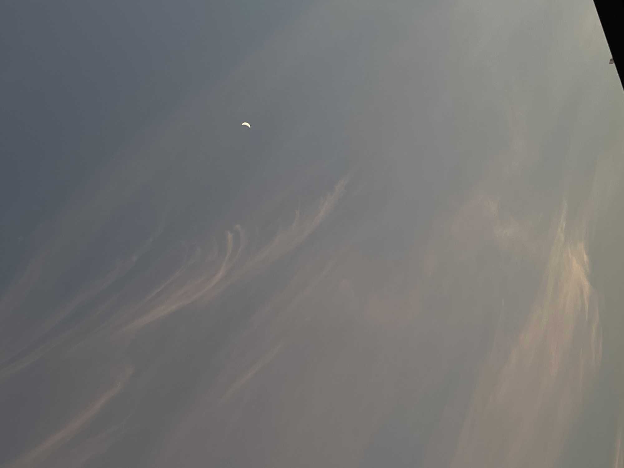 This is a vertical, eye-level, full-shot photograph of a dusky sky with wispy clouds and a crescent moon. The sky is a muted blue-gray color, transitioning to lighter tones towards the bottom of the frame, suggesting the last light of day. Delicate, light pink and white clouds are streaked across the upper left portion of the sky, appearing thin and ethereal. A small, bright crescent moon is visible in the upper right quadrant, casting a subtle glow. The bottom edge of the image is occupied by the dark silhouette of a building or rooftop, suggesting an urban or suburban setting. The location in Sayama, Japan is not discernible from the image itself.