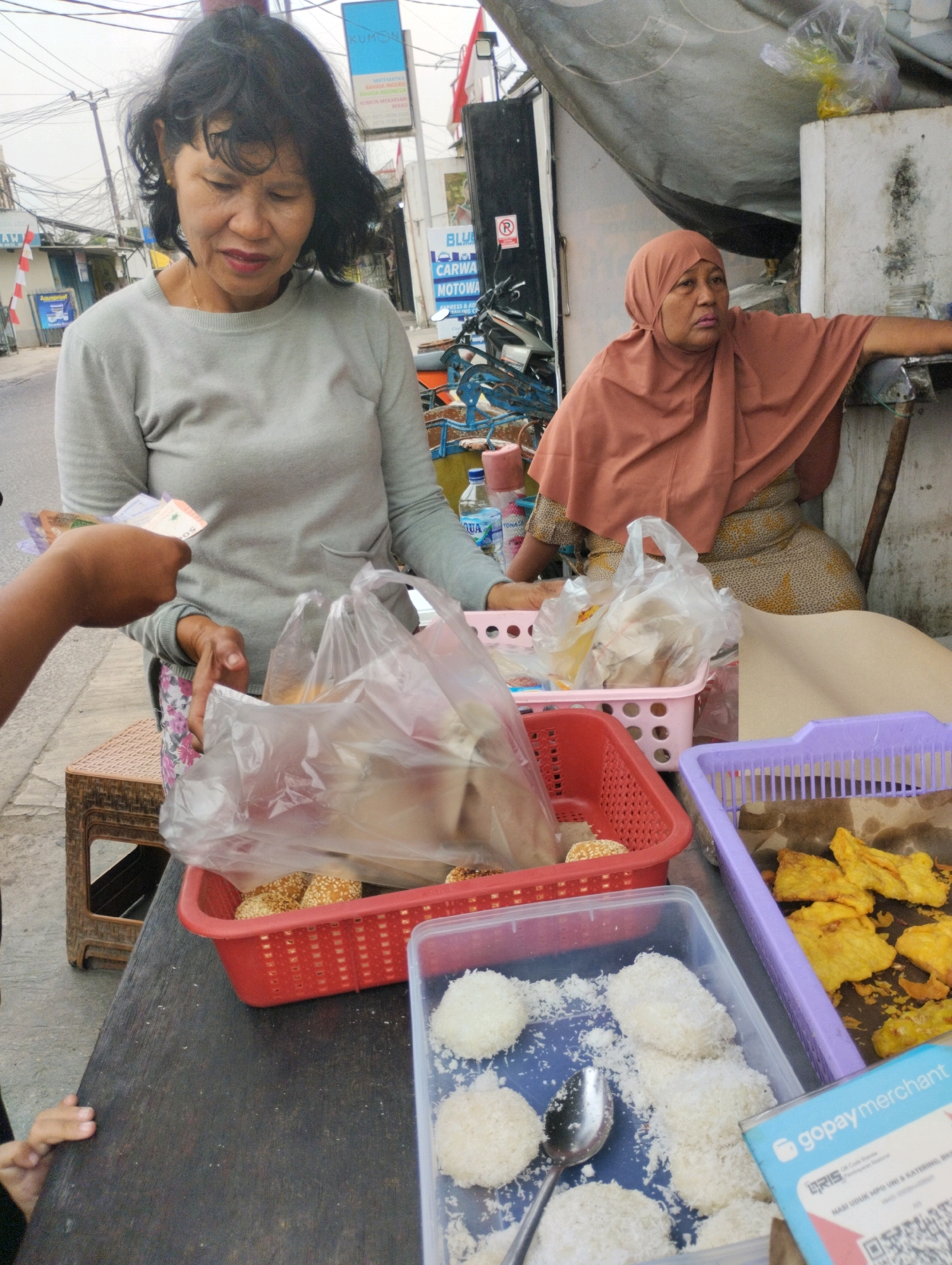 In Bekasi, Indonesia, a street vendor, a woman with dark, shoulder-length hair and wearing a grey sweater, is in the process of a transaction. She is holding a bunch of Indonesian Rupiah banknotes and appears to be handing them to someone or receiving them. In front of her, on a dark wooden surface, are several red and purple plastic baskets containing food items. The red basket holds round, sesame-seed-covered pastries, while the purple basket contains fried food, likely tempeh. A clear plastic container nearby displays more round pastries coated in shredded coconut, with a spoon resting among them. To the right, another woman, wearing a brown hijab and a patterned dress, leans against a white pillar, with her arm resting on a metal pole. Behind the vendor, a blue sign with text is visible, and further back, electrical wires and poles indicate an urban setting. The ground is paved, suggesting a street or sidewalk environment.