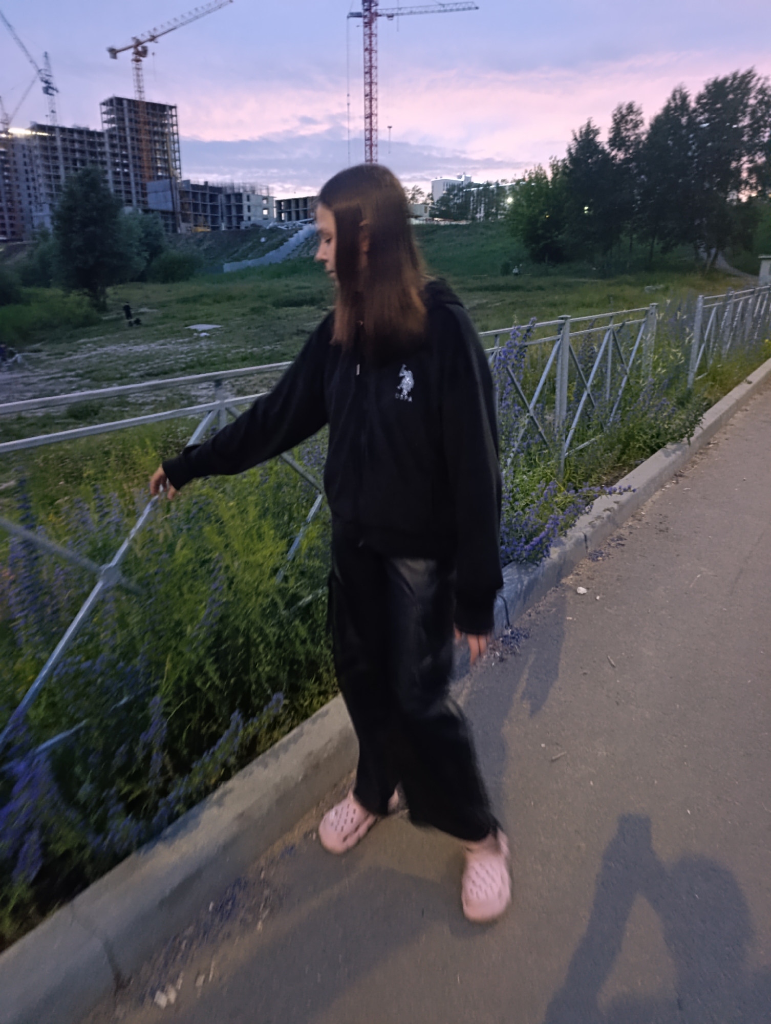 A young woman with long brown hair, wearing a black zip-up hoodie and black pants, stands on a paved surface next to a metal fence. She is touching the fence with her right hand. Pink clogs are visible on her feet. Beyond the fence, there is an area of green vegetation with purple flowers. In the background, construction cranes and unfinished buildings are silhouetted against a twilight sky. The scene appears to be an urban environment, possibly a construction site. The presence of construction cranes and unfinished buildings suggests an area undergoing development. The lighting indicates it is either dawn or dusk. The overall impression is one of an outdoor, public space in a city.