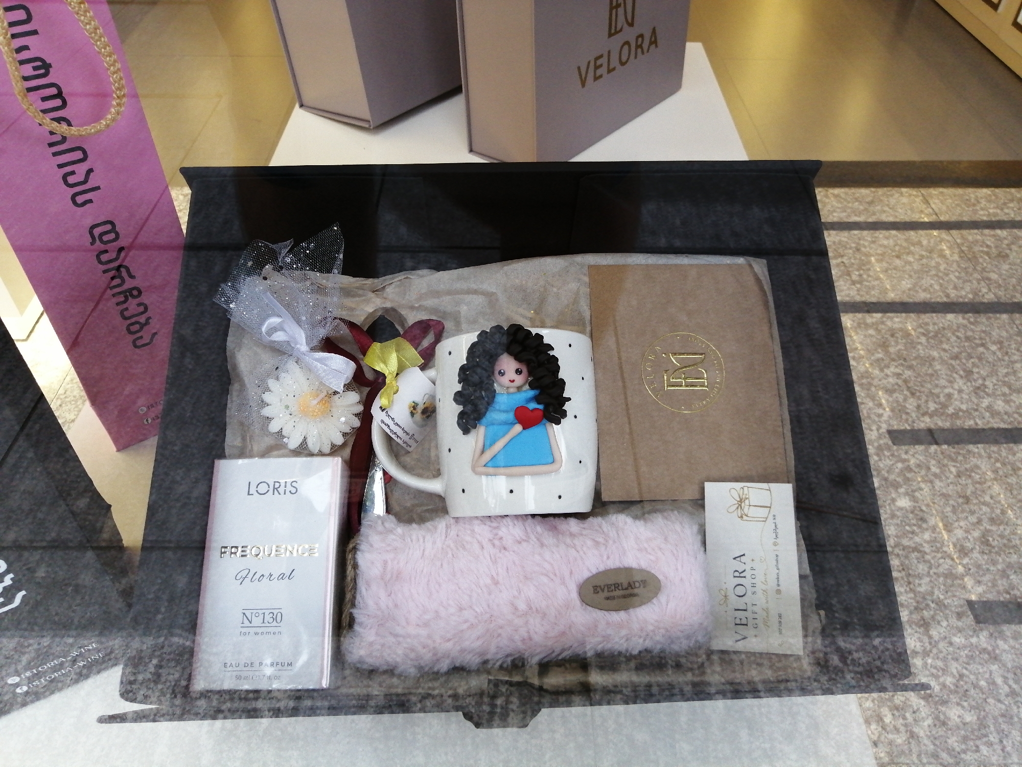 The image shows a gift box displayed in what appears to be a shop window. The gift box contains several items including a white mug with a decorative figure on it, a small pink furry pouch, a perfume bottle, and a card. The mug has a drawing of a person with dark curly hair, wearing a blue dress and holding a red heart. The perfume bottle is labeled "LORIS FREQUENCE Floral N°130 for women". The pink furry pouch has a label that reads "EVERLADY". There is also a brown card with gold lettering that says "VELORA GIFT SHOP". The background suggests an indoor environment, possibly a retail space, with tiling on the floor and a glimpse of a display behind the gift. The overall impression is a curated gift set, likely for sale. The location is identified as Batumi, Georgia, implying this shop is situated within that city.
