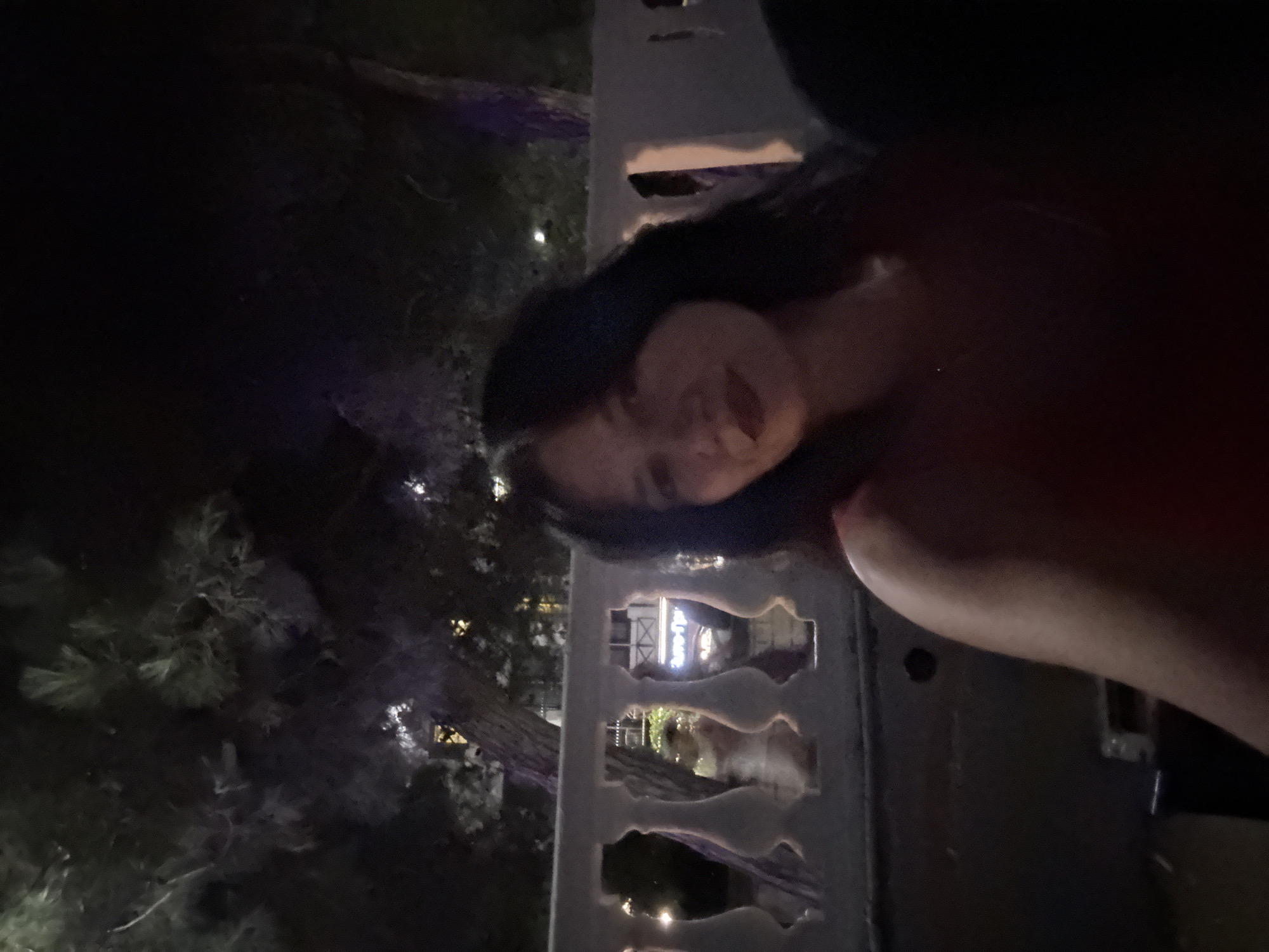 A woman with dark hair and red lipstick is looking at the camera at night. She is wearing a red top. She is seated in front of a white ornate balustrade. Behind her, there are trees and buildings with lights on. The overall lighting is dim, suggesting it is nighttime or a very dark indoor setting.