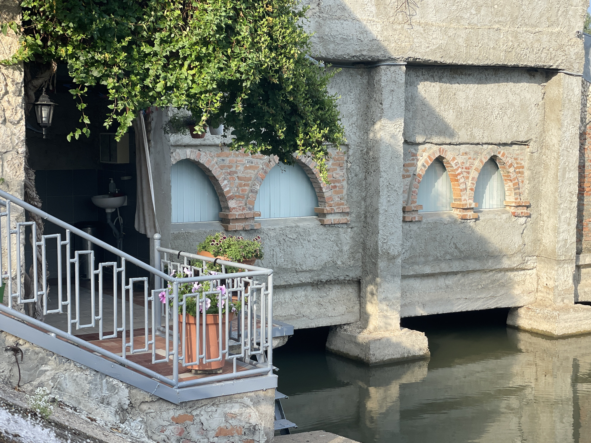 In the city of Colibași, Moldova, a waterside structure features a grey metal railing and stairs leading down to water. Potted plants with pink and purple flowers are placed on the railing. The building itself is made of rough concrete and red brick, with arched windows covered by pale blue doors. Overhead, green foliage drapes from the upper part of the structure. The water reflects the building and the sky. To the left, an outdoor sink and a hanging lamp are visible in an alcove. The overall scene depicts a rustic, waterside architectural setting.