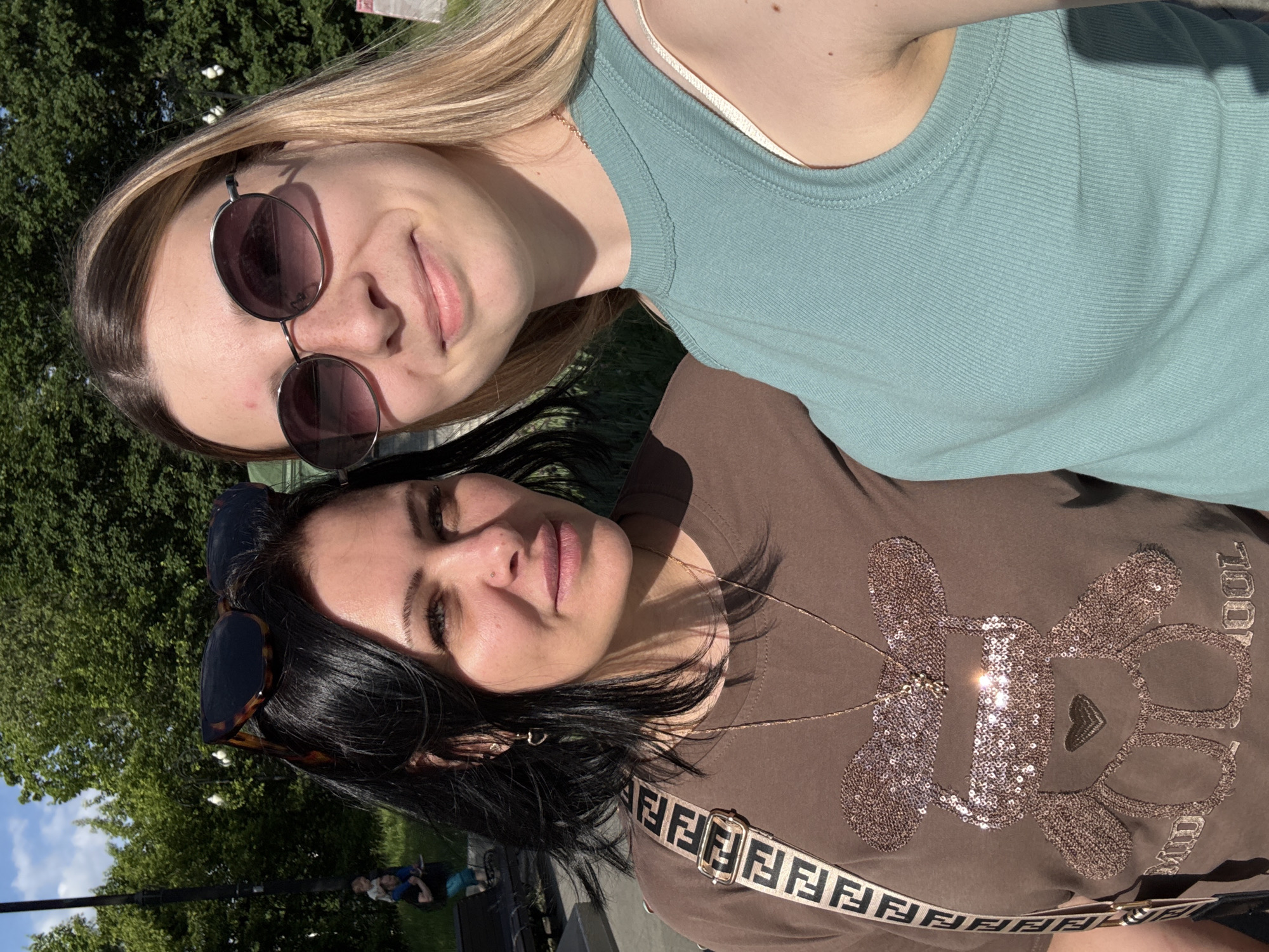 The image shows a rotated close-up selfie of two women posing outdoors on a sunny day, likely in a park in Moscow, Russia.
The woman positioned at the top of the frame has long blonde hair and is wearing round, dark-lensed sunglasses. She is dressed in a light blue ribbed tank top and has a pleasant, smiling expression.
The woman at the bottom of the frame has dark hair, with tortoiseshell sunglasses pushed up on her head. She is wearing a brown t-shirt featuring a large, sparkling sequined design that resembles a teddy bear-like character with glasses, a small heart, and the word "COOL" written vertically on the right side. A gold chain necklace is visible, and a strap with the repeated "FENDI" logo runs across her chest. She maintains a neutral expression while looking towards the camera.
The background consists of lush green trees and a clear blue sky with some white clouds, indicating bright daylight. The strong sunlight creates visible highlights and shadows on the subjects.