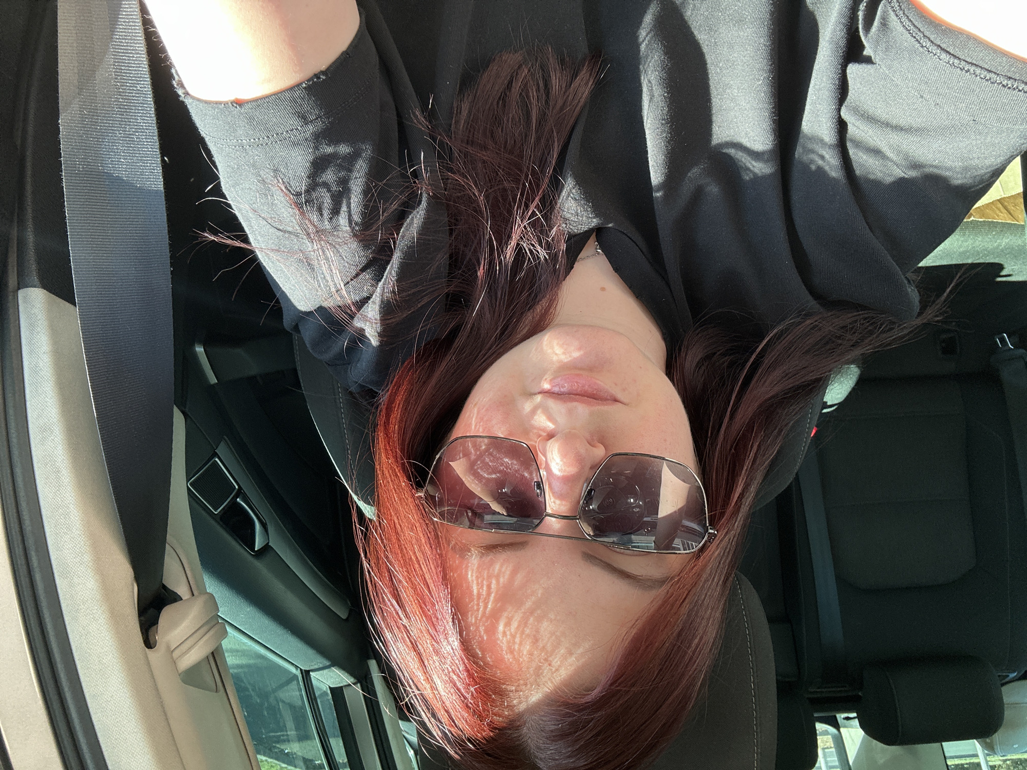 A medium shot captures a young woman with auburn hair, wearing sunglasses and a black t-shirt, inside a car. She is seated in the driver's seat, with the passenger seat and a portion of the back seat visible behind her. Sunlight casts dappled shadows across her face and clothing, suggesting it is daytime. The interior of the car appears to be standard, with dark fabric seats and a seatbelt prominently displayed to her right. The background hints at an urban environment with some greenery visible through the car windows, possibly indicating a street or park setting in the City of Zagreb, Croatia.