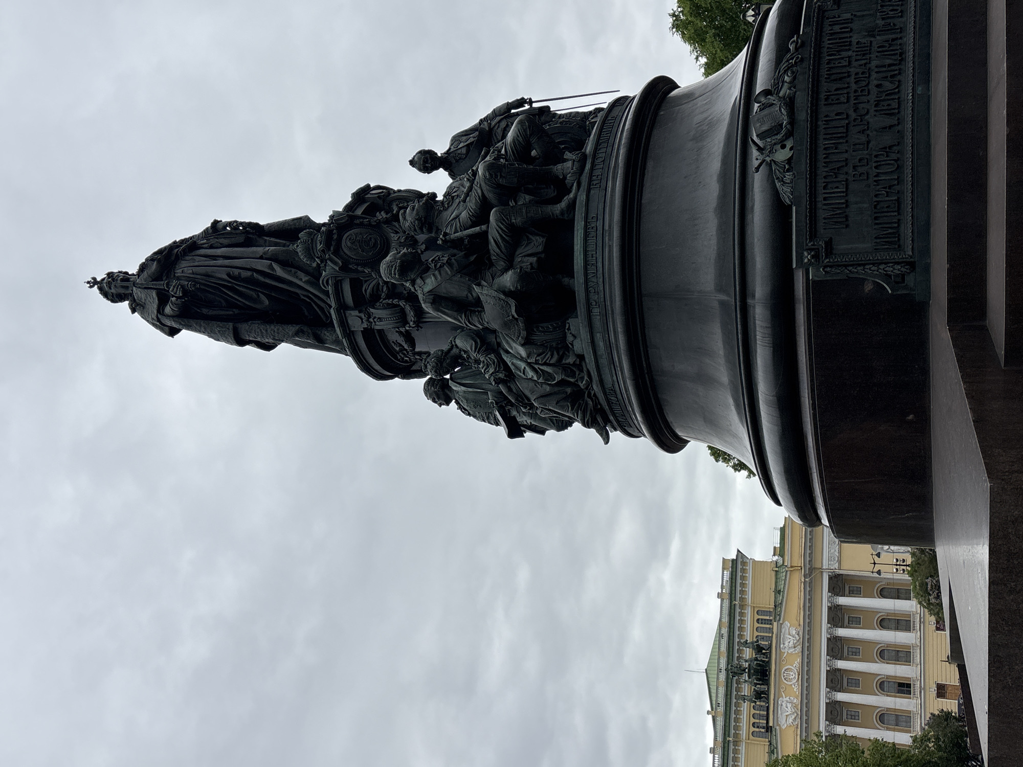 The image shows the Monument to Catherine the Great in Saint Petersburg, Russia. The monument depicts Catherine the Great standing at the top, surrounded by figures of notable contemporaries and advisors. The monument is made of bronze and stands on a granite pedestal. In the background, the facade of a classical building is visible. The sky is overcast.