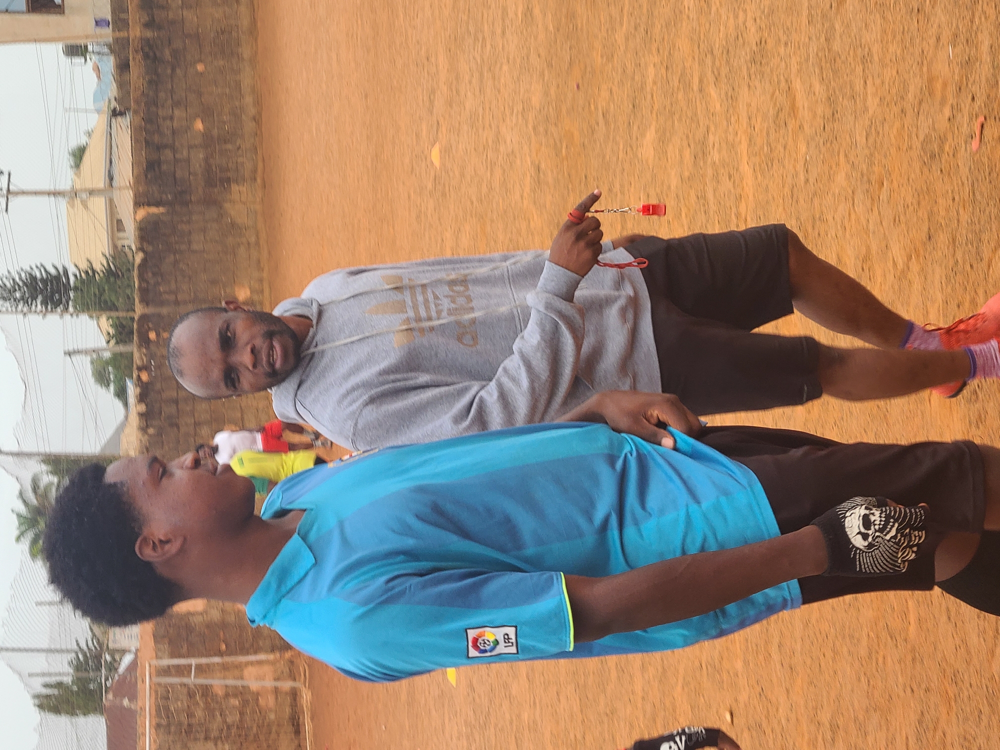 Two men are outdoors. The man in the foreground wears a blue shirt with a badge that reads "LFP," black shorts, and skull-patterned gloves. Behind him, a man in a grey Adidas hoodie and black shorts holds a red whistle. The setting appears to be an open space with a brick wall and foliage in the background. The location is the city of Unknown, Unknown.