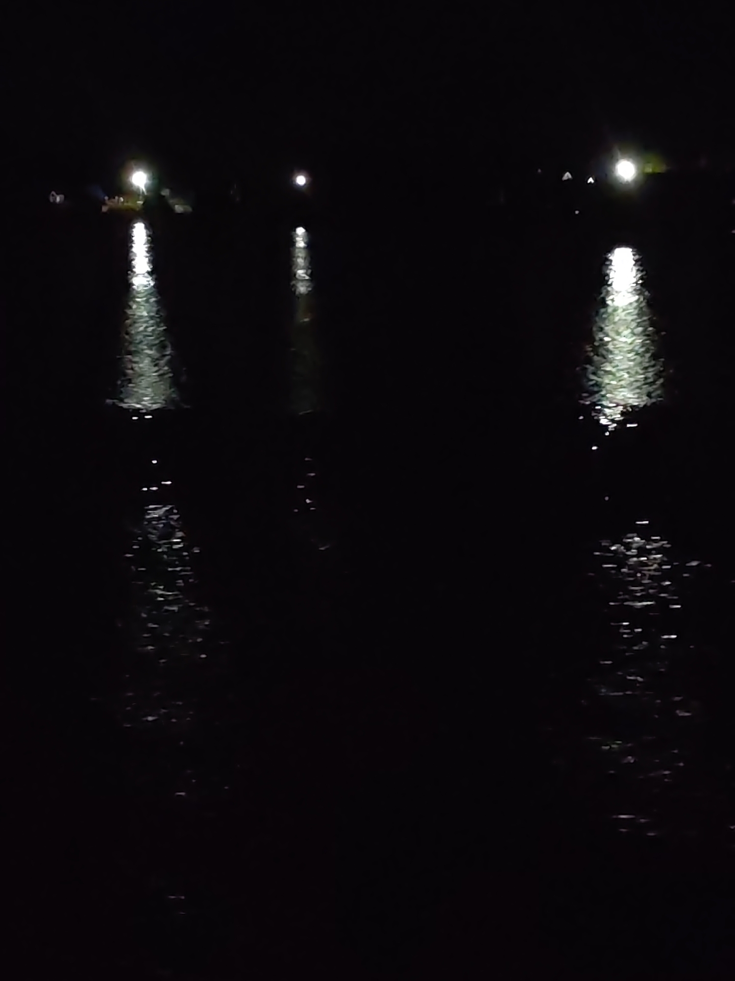 The image appears to depict a body of water, likely a lake or river, at night. There are three distinct light sources visible on the opposite shore, their reflections stretching across the water. The water's surface is disturbed, causing the light reflections to break up and scatter. The rest of the scene is dark, making it difficult to discern any further details about the surrounding environment. There is no information available about the location in the city of Zvoz, Russia.