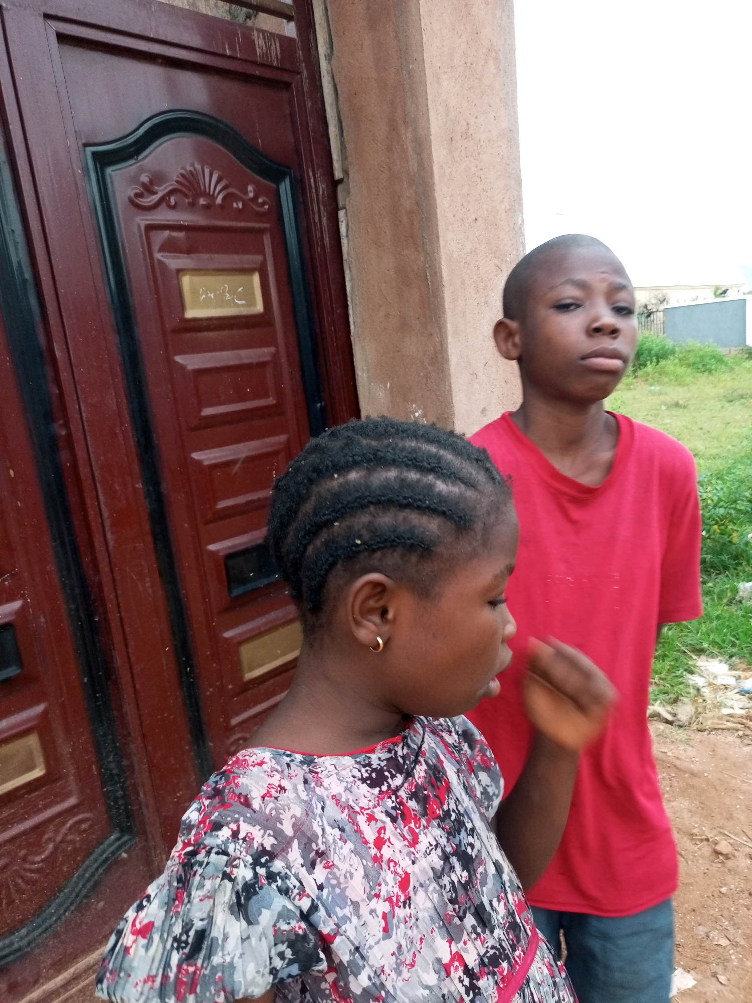 Two young people, a girl in the foreground and a boy behind her, stand in front of a dark red double door. The girl, facing left, has her hair braided and wears a colorful dress with a floral pattern. She has a gold hoop earring in her left ear and her right hand is raised towards her face. The boy, facing forward and slightly to the right, has a shaved head and wears a red t-shirt and blue jeans. He is looking towards the camera. The door is ornate, with raised panels and a golden plaque with an inscription. Behind the children, there is a sandy ground and some green vegetation, suggesting an outdoor setting. The architecture and vegetation are consistent with a location in Tempure, Nigeria.