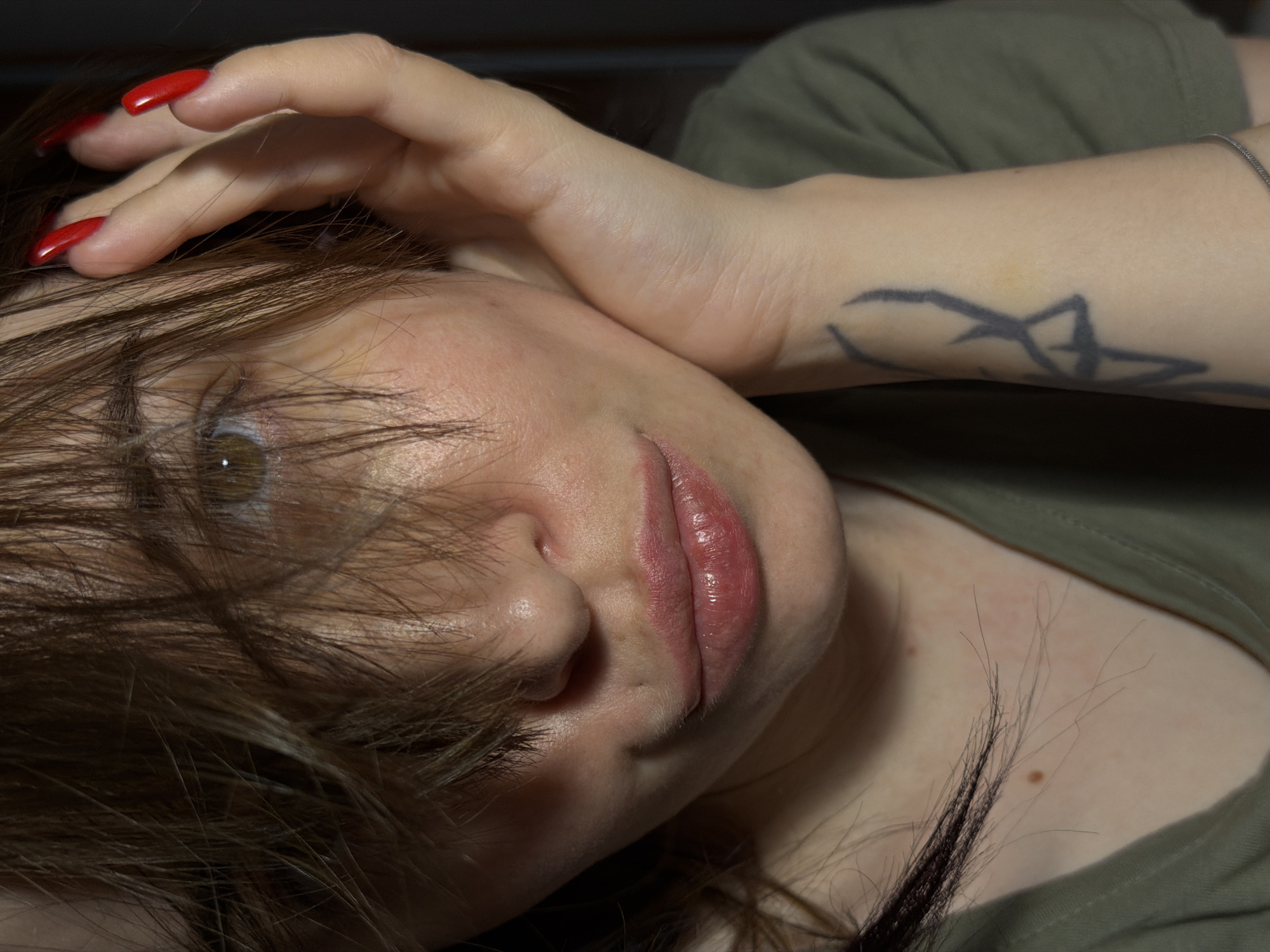 A close-up, head-and-shoulders shot shows a person with brown hair covering their face, obscuring most of their features. Their right hand, with bright red, long, pointed fingernails, is raised to their face, touching their hair. A portion of their left arm is visible, featuring a dark, abstract tattoo. The person is wearing a green t-shirt. The background is dark and indistinct, suggesting an indoor setting. The image is cropped tightly, focusing on the upper body and face.