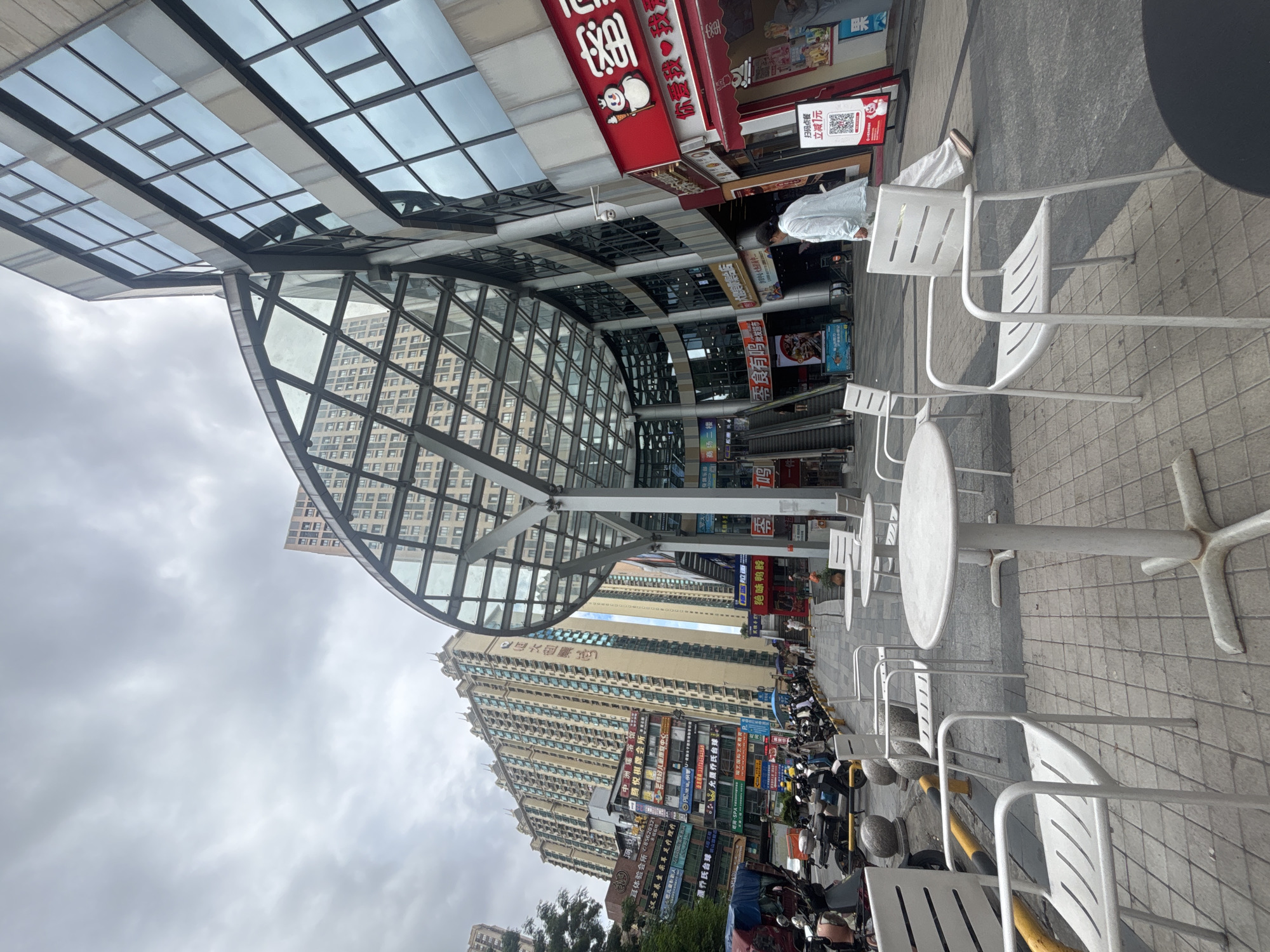 The image depicts an outdoor seating area in front of a modern building with a glass-covered entrance in Nanhai District, China. Several white metal tables and chairs are arranged on a tiled walkway. In the background, a tall, multi-story building with numerous windows dominates the skyline. A street with parked motorcycles and scooters is visible to the left of the seating area, along with a street vendor's stall displaying various items. A person in a light blue outfit walks away from the camera towards the building. The sky is overcast, suggesting a cloudy day. The architecture is a blend of modern commercial design with traditional elements, as indicated by the signage and the overall urban setting.