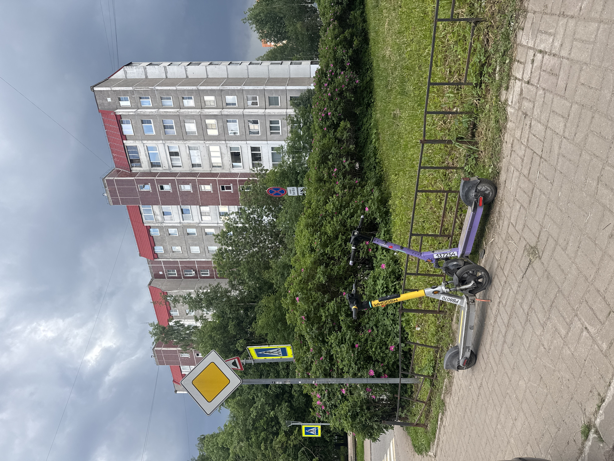A cluster of electric scooters is parked on a brick sidewalk in front of a large, multi-story apartment building with a red roof. Two scooters are prominent, one yellow and white with "WHOOSH PB8070" printed on it, and the other purple with "137254" printed on it. Behind the scooters is a dense green bush with pink flowers, and beyond that, a grassy area bordered by a metal fence. The apartment building has many windows and a mix of concrete and red brick facades. To the left of the building, a street sign with a yellow diamond and black border, along with other road signs, are visible. The sky above is cloudy and gray, suggesting overcast weather. The environment appears to be a residential urban area.