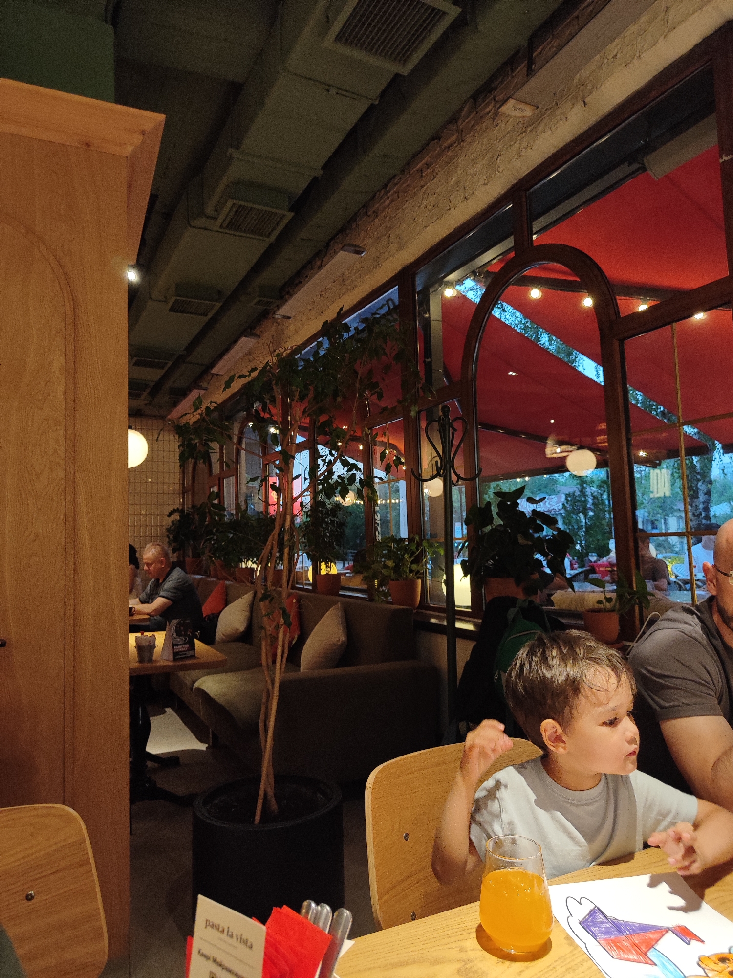 The image depicts an indoor scene, likely a restaurant or cafe, with a child sitting at a table in the foreground. The child is wearing a light blue t-shirt and has a glass of orange juice in front of them, along with a drawing on a piece of paper. In the background, there are other patrons seated at tables, and the restaurant's interior features wooden furniture, plants, and large windows that offer a view of the outdoors. The ambiance suggests a relaxed dining or social setting. The question about the location in Almaty, Kazakhstan cannot be answered from the image alone.