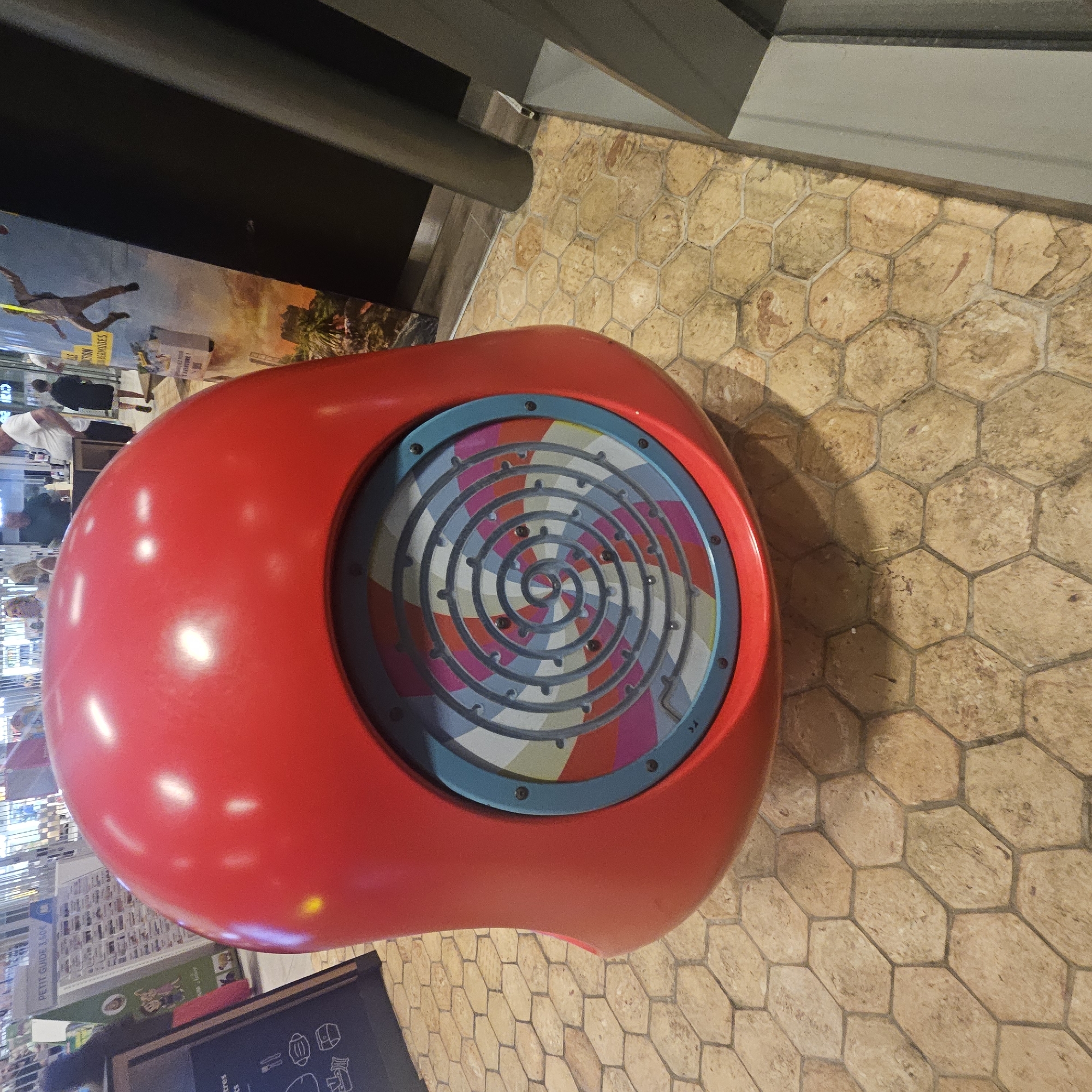 A large, red, spherical object, likely a piece of playground equipment or a decorative installation, is situated on a tiled floor. The object has a circular panel on its front, featuring a colorful maze with small dark balls. The surrounding environment appears to be an indoor public space, possibly a shopping mall or a leisure center, with posters and people visible in the background. The floor is covered in hexagonal tiles with a textured, light brown color. The image is taken from a low angle, looking up at the red object. The overall lighting suggests an indoor setting with artificial illumination. The location is Lançon-Provence, France.