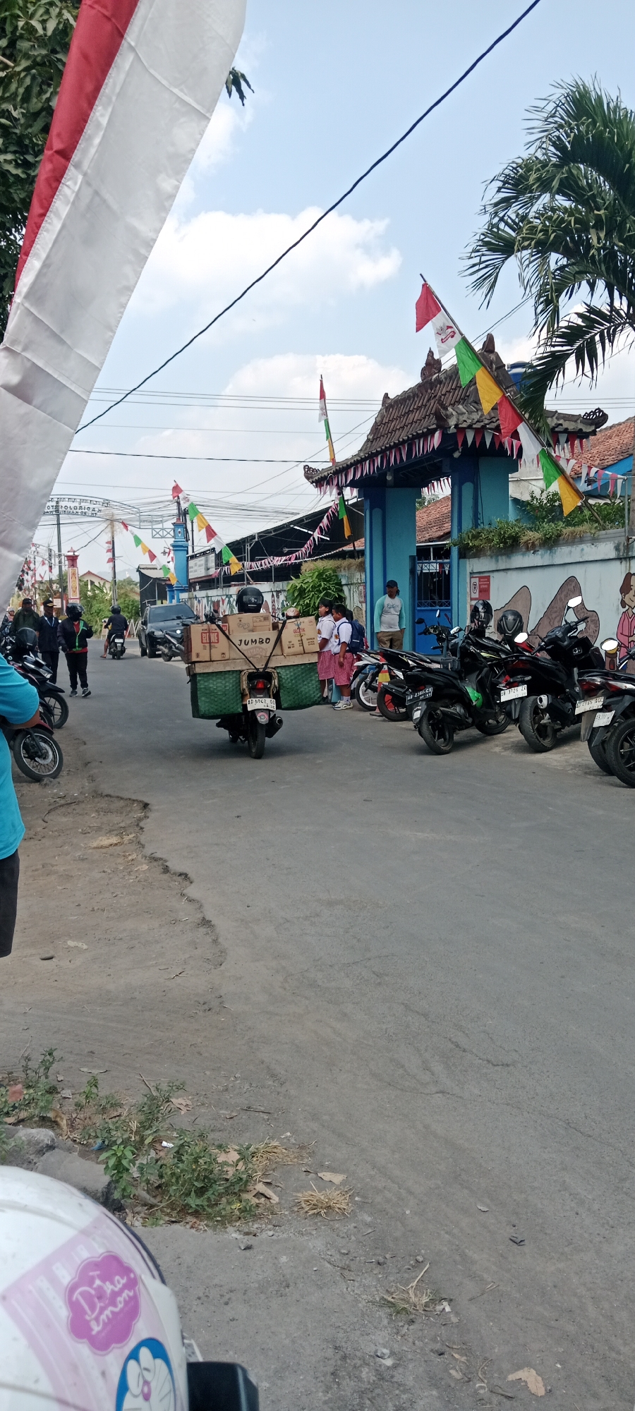 The image depicts a street scene in Purwomartani, Indonesia, likely on a bright, cloudy day. In the foreground, a white motorcycle helmet with stickers is partially visible. The road surface shows signs of wear and damage, with a large pothole on the left side. A motorcycle carrying a large, green basket filled with cardboard boxes is centrally positioned. The boxes are labeled with "JUMBO" and "KARTON." Two school-aged girls in white shirts and red skirts, carrying backpacks, stand near the motorcycle.
Behind them, a blue gate with decorative roofing and banners is visible, suggesting a public building or school entrance. Several motorcycles are parked to the right of the gate. The background shows power lines, trees, and buildings. Indonesian flags and colorful triangular pennants are strung across the street, indicating a possible celebration or national event. People are seen walking and standing around the area. The overall atmosphere appears to be that of a typical local street in a residential or semi-commercial area.