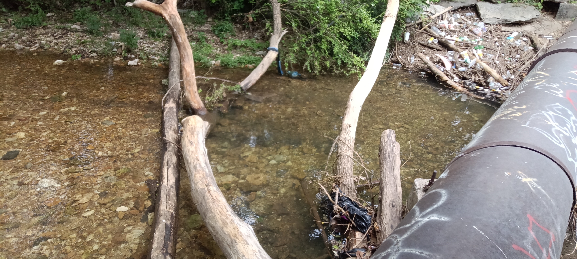 The image shows a stream or creek bed in a natural setting, likely located in or near Tahlequah, Oklahoma. The water is shallow and clear, revealing a rocky bottom. Several fallen tree limbs and branches are scattered across the streambed, creating natural obstacles. A large metal pipe, covered in graffiti, runs along the edge of the stream. Debris, including plastic bottles and other litter, is concentrated on one side of the creek near the pipe, indicating a pollution issue. The banks of the creek are lined with vegetation, including trees and bushes. The overall scene suggests a natural environment impacted by human activity.
