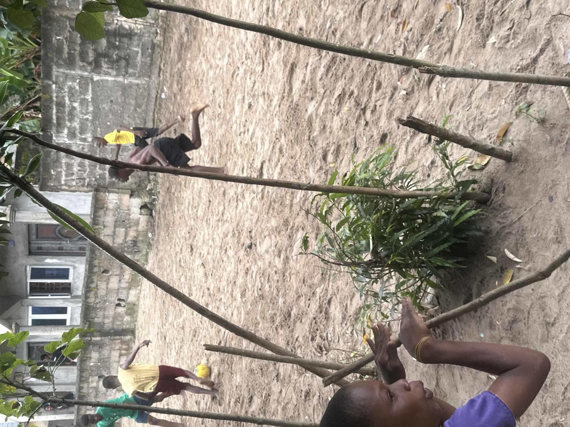 A low-angle, medium shot captures a group of young boys playing soccer on a sandy field in Badagry, Nigeria. In the foreground, a boy wearing a purple shirt is partially visible on the left, looking towards the action with his hands outstretched. The sand is uneven, with small plants and debris scattered across its surface. In the mid-ground, two boys are engaged in the game. One boy, wearing a yellow shirt and red shorts, is kicking a yellow soccer ball. Another boy, wearing black shorts, is running towards the ball. In the background, a brick wall forms a boundary, with a building visible above it. The sky is overcast, suggesting a cloudy day. The scene is dynamic, conveying the energy and excitement of the game.