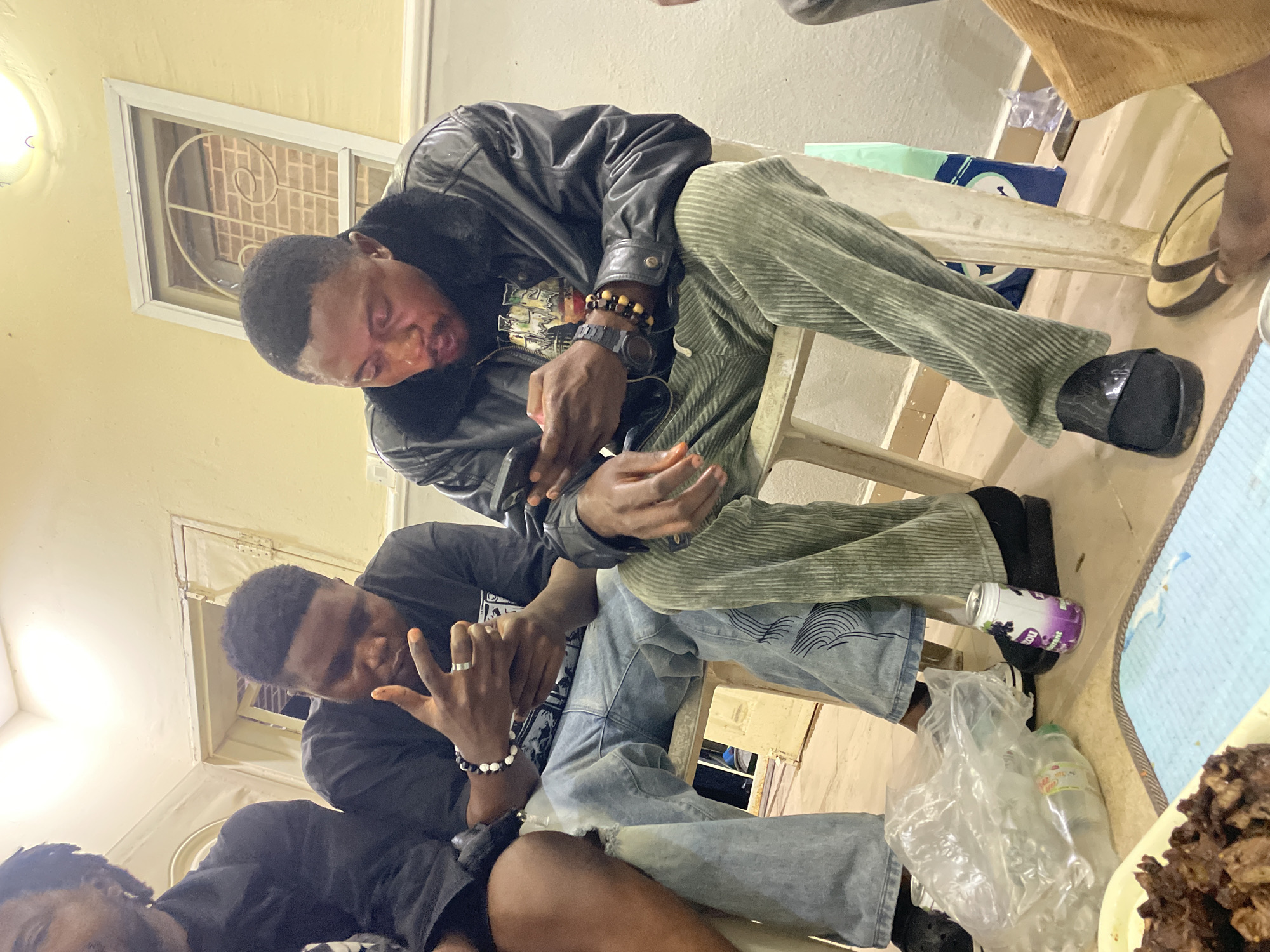 Three Black men are sitting in a room. One man is wearing a black leather jacket and sage corduroy pants. Another is wearing a black T-shirt and blue jeans. The men are seated on white plastic chairs. There is food and drinks on a table to the right. The location is possibly Jos, Nigeria.