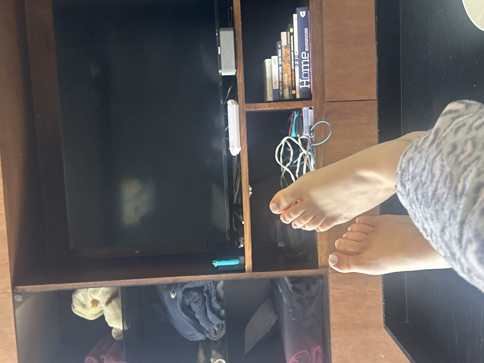 The image displays an indoor scene, consistent with a residential living area in Huixquilucan de Degollado, Mexico. The perspective is low-angle, looking towards an entertainment setup.
In the immediate foreground, a person's bare feet are prominently featured, elevated and resting. The toenails on both feet are painted with a light-colored polish, likely white or very pale pink. The individual's lower legs are partially obscured by patterned loungewear or sweatpants in shades of gray and white.
The background is dominated by a dark wooden entertainment unit or shelving system. A large, dark-screened television is centrally positioned on an upper shelf of this unit; a faint, undefined reflection is visible on its surface. Directly beneath the television, on a wooden shelf, several objects are arranged: a white rectangular remote control is on the left, next to a blue/turquoise Nintendo Switch Joy-Con controller amidst a cluster of white and blue cables. Further right on this shelf is a gray rectangular speaker or soundbar.
To the left of the central television, the shelving unit contains three visible compartments:
- The top-left compartment holds a light-colored stuffed animal, possibly a bear, with a visible orange-brown marking, situated next to folded dark red towels.
- The middle-left compartment contains neatly folded blue towels.
- The bottom-left compartment displays folded gray and pink textiles.
To the right, on a lower shelf of the entertainment unit, a stack of books is visible. The spines of multiple books are legible, with the lowest and largest book clearly displaying the title "Home."
The scene conveys a casual, domestic environment.
