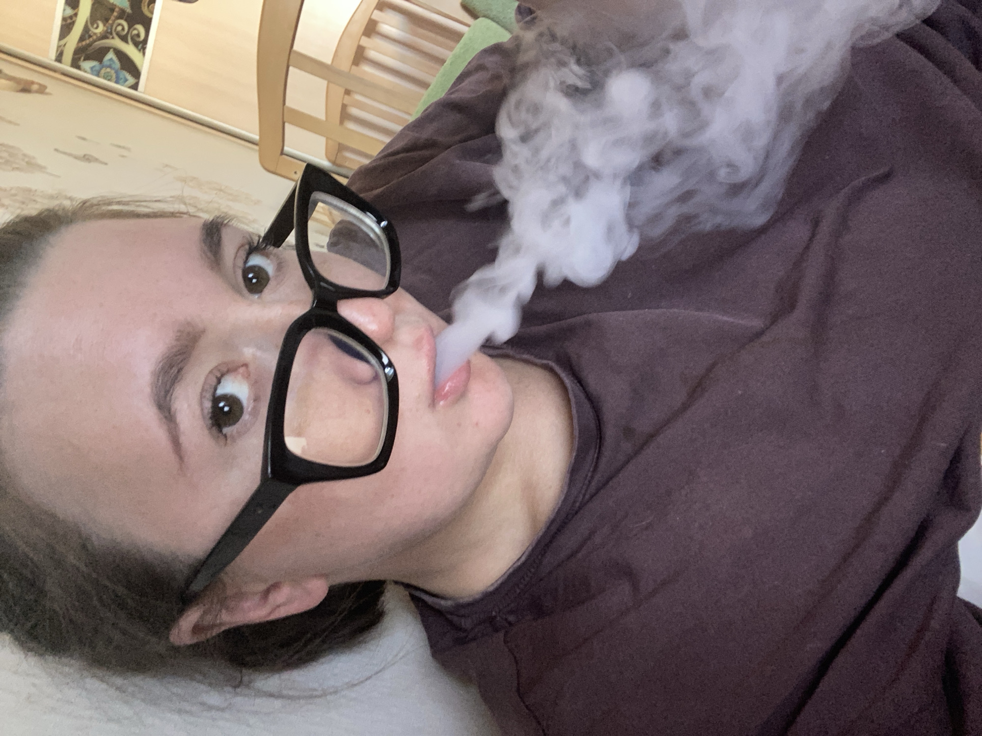 A young woman with brown hair and large black-rimmed glasses is lying down and exhaling a cloud of white vapor, suggesting she is vaping. The image is a close-up of her face and upper body, with the background blurred. She is wearing a dark brown long-sleeved shirt. Her eyes are open and looking towards the camera, and her lips are slightly parted. The lighting suggests it is daytime, possibly indoors. The background includes a hint of wooden furniture, possibly a crib or chair, and some patterned wallpaper or tapestry. The overall mood is casual and relaxed. There is no visible text in the image. The location context provided is Chaikovsky, Russia.