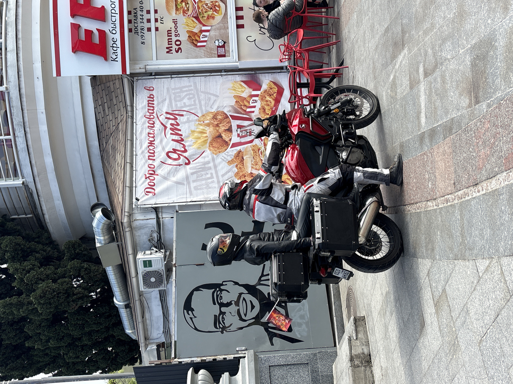 Here's a detailed description of the image:
**Location:** This scene is in город Ялта, Ukraine. This is indicated by the sign "Добро пожаловать в Ялту" (Welcome to Yalta).
**Subjects:**
* Two individuals are sitting on a motorcycle. Both are wearing helmets and riding gear.
**Objects:**
* A red and black motorcycle is parked on a sidewalk. It has storage boxes attached to the rear.
* A KFC (or similar fast-food restaurant) is visible in the background. There are KFC advertisements featuring fried chicken and other menu items.
* Signs with Russian text are visible on the building.
* Red chairs are placed outside the establishment, presumably for outdoor seating.
* A metal ventilation system runs along the side of the building.
* An air conditioning unit is attached to the building.
**Actions:**
* The two individuals are sitting on the motorcycle, possibly having stopped at the KFC.
**Environment:**
* The scene appears to be a street or sidewalk next to a commercial building.
* The weather seems to be sunny, based on the lighting and shadows.
* The sidewalk is paved with gray stone tiles.
* A tree is to the left of the building.