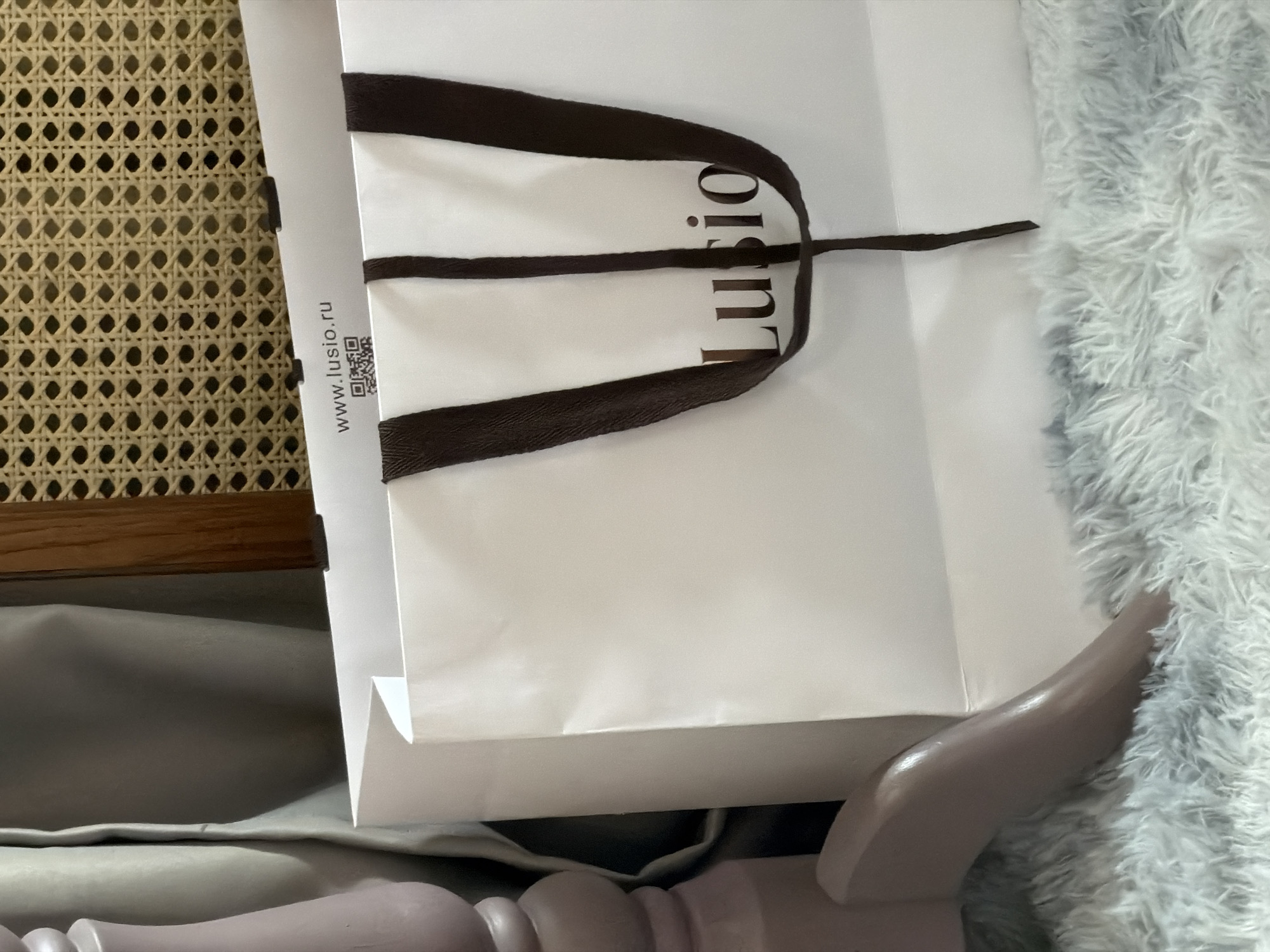 The image depicts a white shopping bag with brown handles and the word "Lusio" printed on it. The bag is placed on what appears to be a piece of furniture with a wicker back. To the right of the bag is a light blue, fluffy material, likely a blanket or rug. The bag also shows the website address "www.lusio.ru" and a QR code. Given the ".ru" domain and the brand name "Lusio," it is reasonable to infer that the scene could be located in Moscow, Russia, where Russian businesses operate. The shopping bag sits on a soft, gray-colored surface and rests against a curved, painted element.