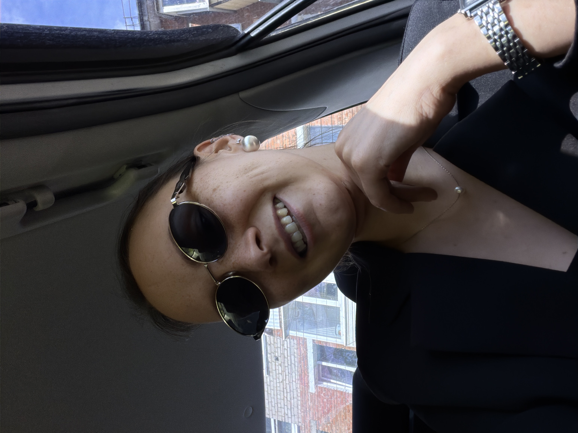 A medium shot captures a woman wearing sunglasses and a black blazer, seated inside a vehicle. Her left hand rests on her chest, near a delicate pearl necklace. On her left wrist, a metallic watch is visible. In the background, brick buildings with windows are seen, suggesting an urban setting. The sky is blue with some clouds. The image is taken from a low angle, looking up towards the woman. The lighting suggests it is daytime.