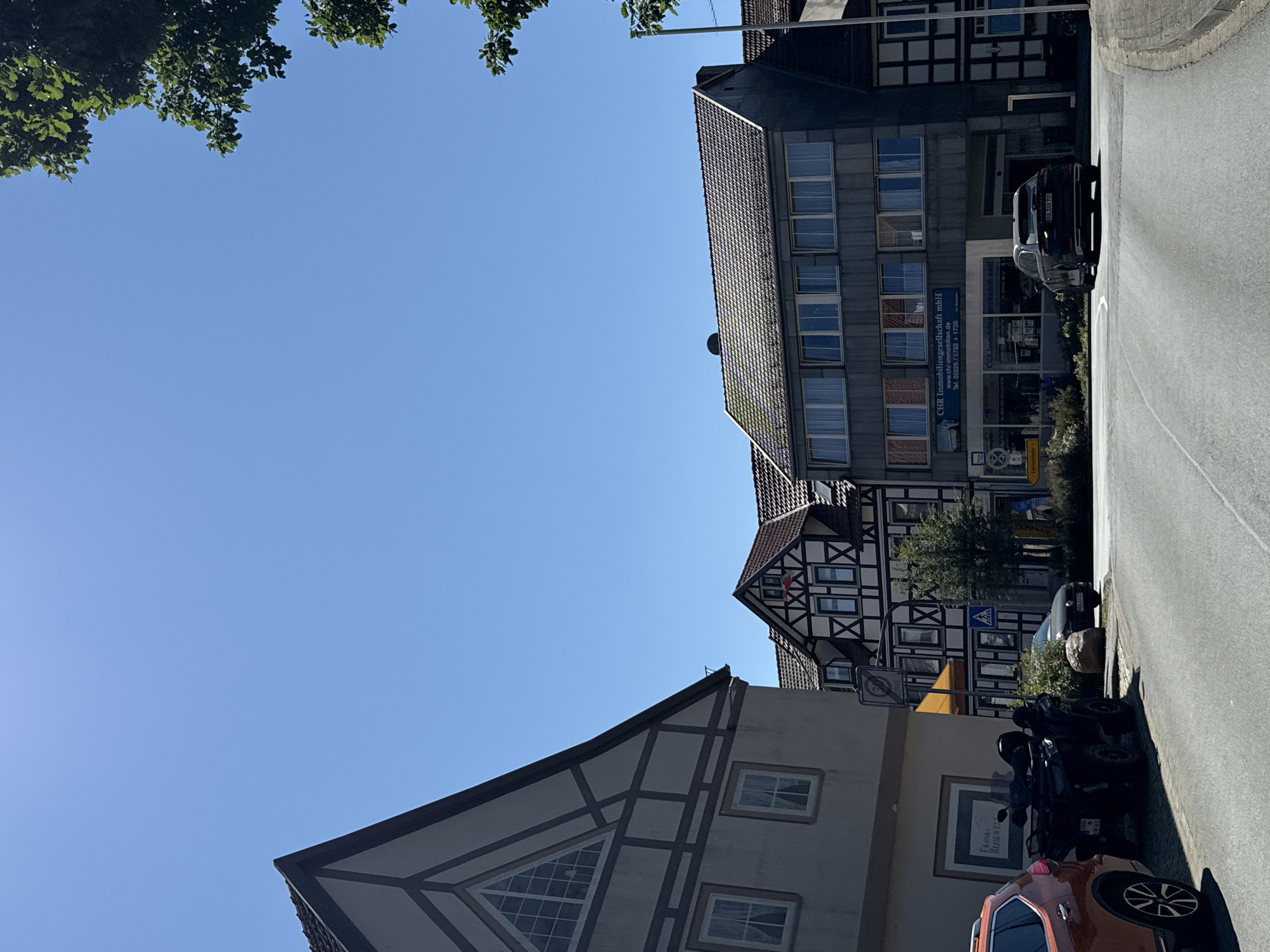 The image captures a street scene in Bad Sachsa, Germany, on a bright, sunny day. In the foreground, a paved road curves to the right. To the left of the road, an orange SUV is parked, and beside it, a black all-terrain vehicle (ATV) is also parked. The building directly behind the vehicles has a light-colored facade with dark-trimmed windows. Further down the street, a building with distinctive half-timbered architecture is visible, featuring white walls and dark wooden beams. Adjacent to this, a modern, grey, multi-story building stands with rows of windows. A dark-colored car is driving away from the viewer on the street, turning into the curve. Overhead, a clear blue sky dominates the upper portion of the image, with the green branches of a tree visible in the top right corner. Signage is present, including a traffic sign with a circular prohibition symbol and an arrow, and a yellow directional sign. The overall impression is a view of a German town on a clear day.