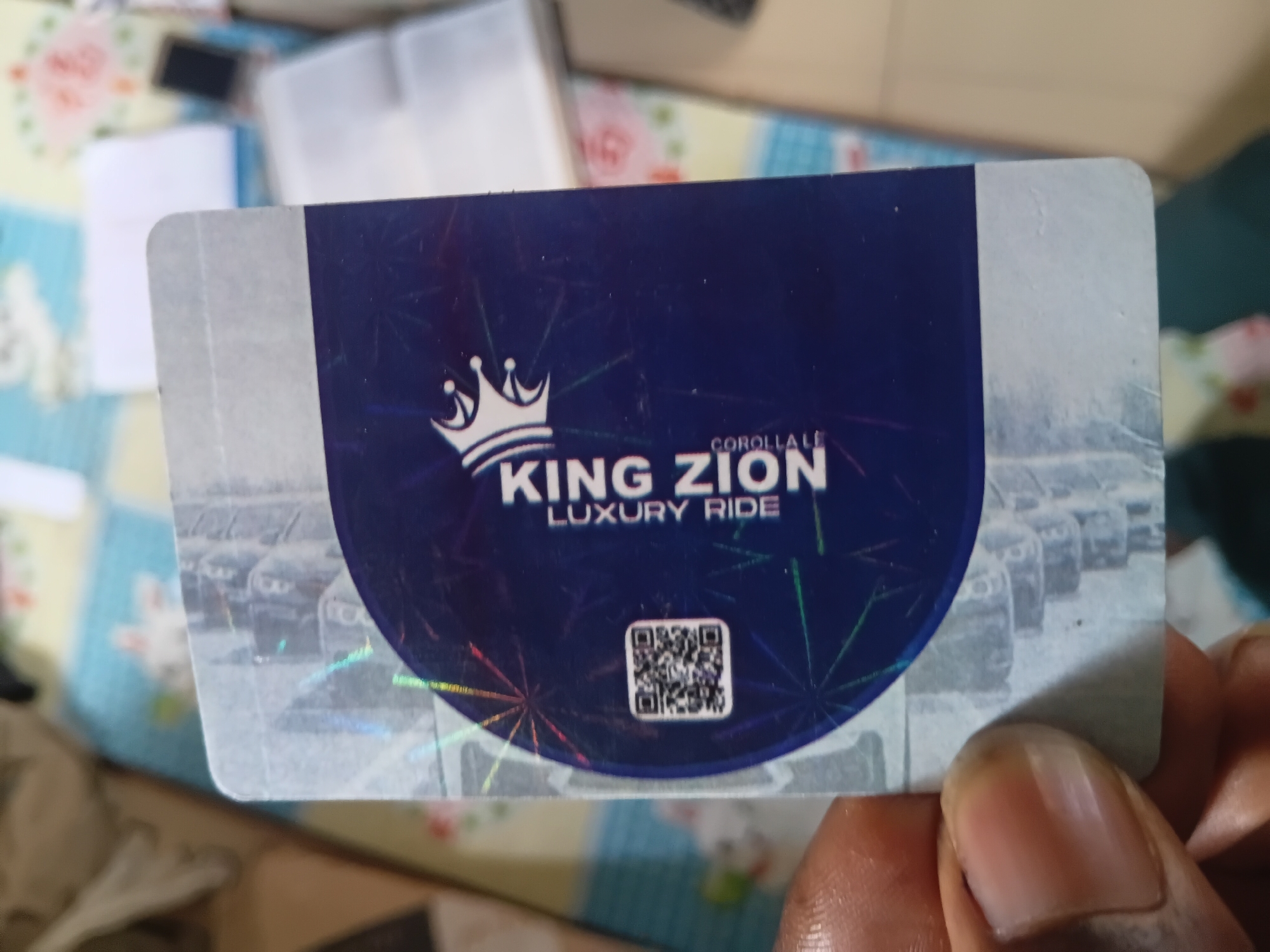 A person's left hand, with a visible thumb, holds a rectangular plastic card with rounded corners against a blurred, light-colored background. The card features a dominant dark blue central section with a white crown logo. Vertically oriented white text on the blue section reads "KING ZION" followed by "LUXURY RIDE" and "COROLLA". A black and white QR code is positioned on the lower left of the blue section. The top and bottom portions of the card are light grey with a faint, indistinct pattern. Holographic elements create rainbow-colored reflections on the card's surface. The background consists of a patterned textile, possibly a bedspread or mat, in light blue, yellow, and white, along with indistinct white papers and a small dark object. The scene is located in Jalingo, Nigeria.