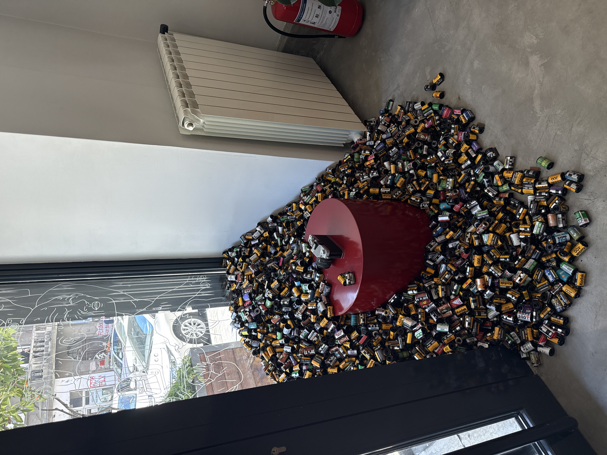 The image depicts an interior space in Belgrade, Serbia. A collection of approximately several hundred film canisters is scattered on the floor around a red cylindrical pedestal, on which a camera sits. A white radiator is mounted on the wall near a fire extinguisher. A window with etched figures provides a view of the street outside. The floor appears to be a gray, polished concrete.