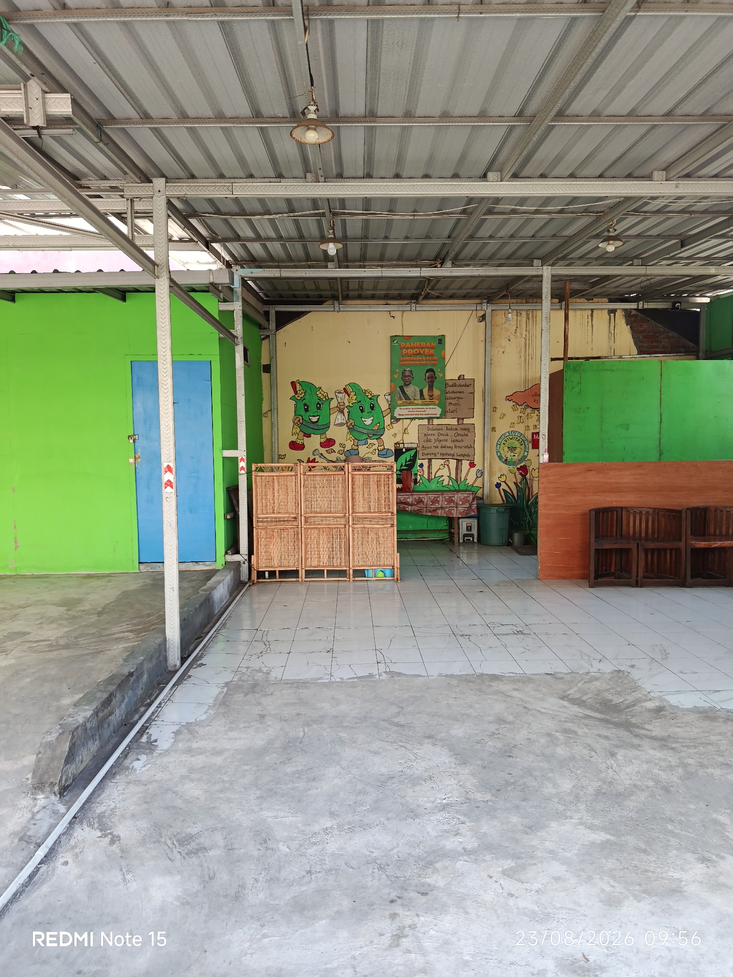 The image depicts an outdoor area with a metal roof supported by metal beams. The floor is paved with tiles and concrete. To the left, a bright green building with a blue door is visible. In the center, there is a set of wooden dividers with woven panels, and to the right, a wall with colorful murals and posters. Several wooden chairs are arranged near a wooden wall on the right side of the scene. The overall environment appears to be a public space, possibly a communal area or a waiting area, located in Keboansikep, Indonesia.