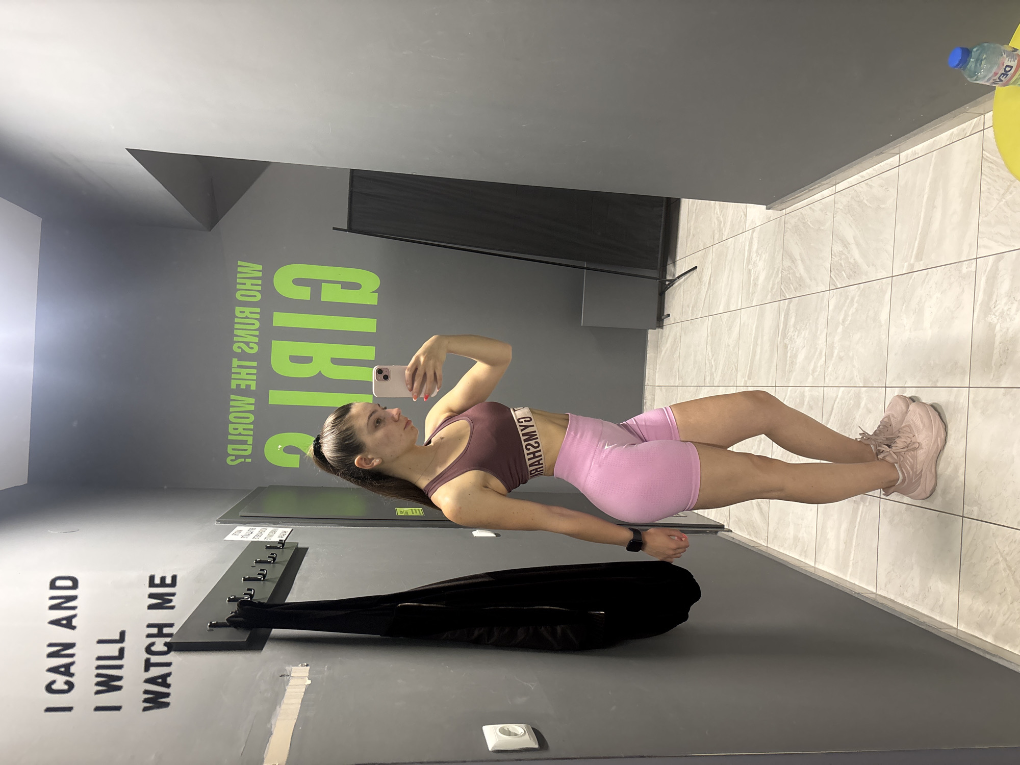 The image shows a young woman taking a selfie in what appears to be a gym changing room in Varna, Bulgaria. The room has grey walls with motivational quotes in English printed on them. The woman is wearing a matching two-piece sportswear set in shades of pink and brown, and pink sneakers. A black jacket hangs on a coat rack behind her. A bottle of water sits on a yellow surface in the background. The floor is tiled in light grey.