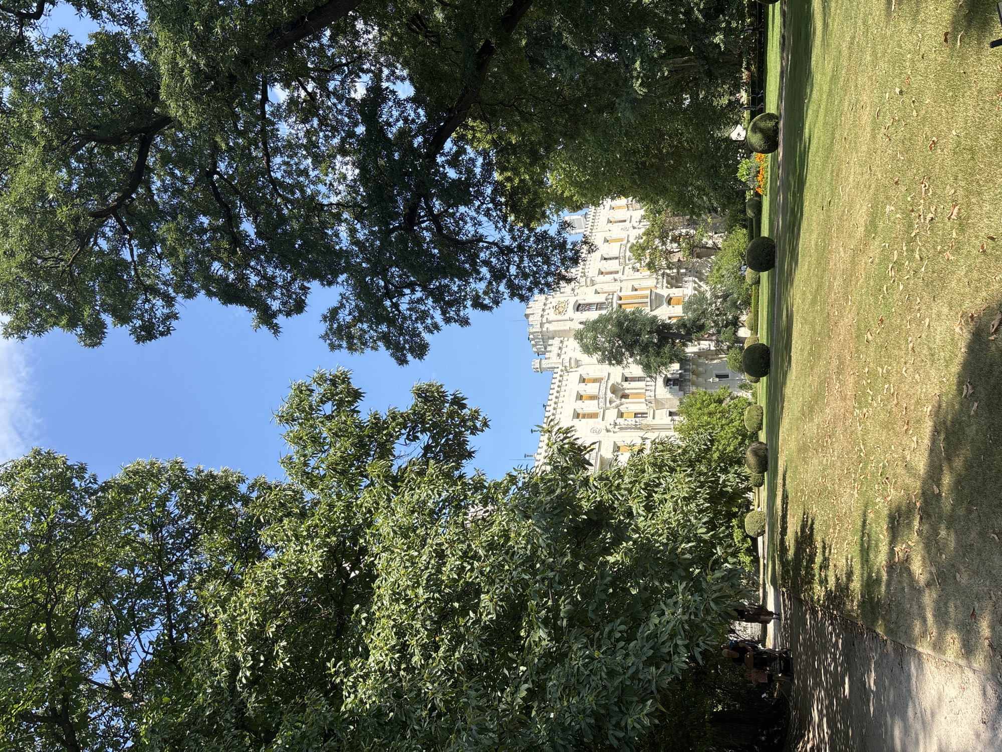 This is a photograph taken in the city of Hluboká nad Vltavou, Czechia, depicting the exterior of Hluboká Castle. The castle, a prominent white structure with numerous windows and architectural embellishments, is visible in the background, partially obscured by lush green trees. In the foreground, a well-maintained lawn with scattered leaves stretches towards the castle. Several round, manicured bushes are dotted across the lawn. On the left side of the image, a paved pathway runs alongside the lawn, shaded by large trees. Two people, one pushing a stroller and another walking, are visible on this pathway. The sky above is a clear, bright blue with a few wispy clouds. The overall scene suggests a sunny day in a park or garden setting adjacent to a historic castle.