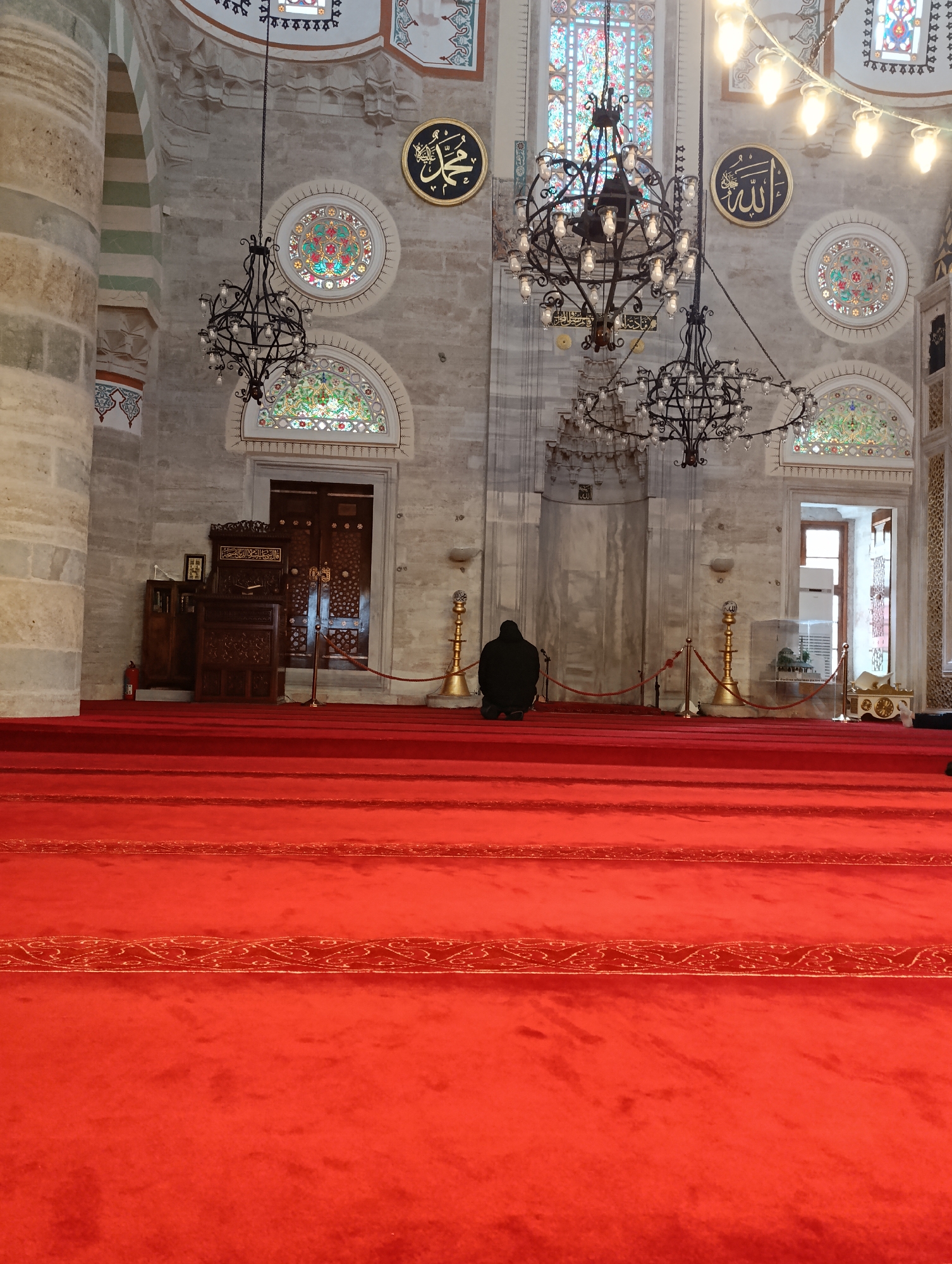 The image depicts the interior of a mosque in Üsküdar, Turkey. A red carpet covers the floor, with lines delineating individual prayer spaces. A person dressed in black is kneeling in prayer. Ornate chandeliers hang from the ceiling, and circular plaques inscribed with Arabic calligraphy are mounted on the walls. Stained-glass windows are visible above the arched doorways. A wooden lectern stands near a closed door on the left. A red rope is held in place by gold colored stands.