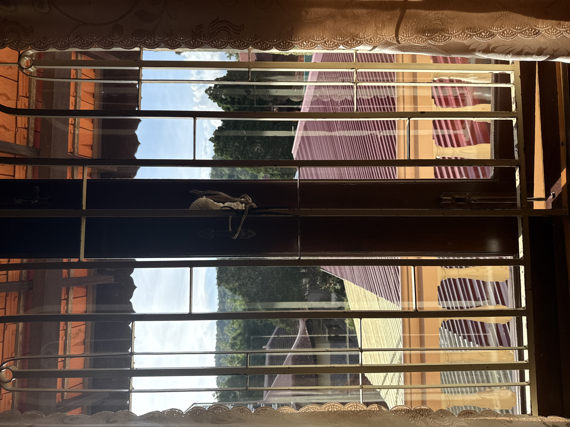 The image is a view through a window with horizontal metal bars and a decorative lace curtain at the top. The window looks out onto a residential scene in Da Lat, Vietnam.
The primary subjects are the reflections and view through the window. The scene outside features a red-tiled roof, some trees, and a pale blue sky with scattered clouds, suggesting it is daytime, likely morning or afternoon. The sunlight is casting shadows and highlights, indicating it is a sunny day.
Notable details include the distinctive corrugated metal roofing, which is common in Vietnam. There are also reflections of what appear to be window blinds or shutters inside the room, adding a layered effect to the image. A small, white object, possibly fabric or a decorative item, hangs from one of the horizontal window bars in the middle section. The overall impression is one of a quiet, domestic scene viewed from within a home. There is no visible text in the image.