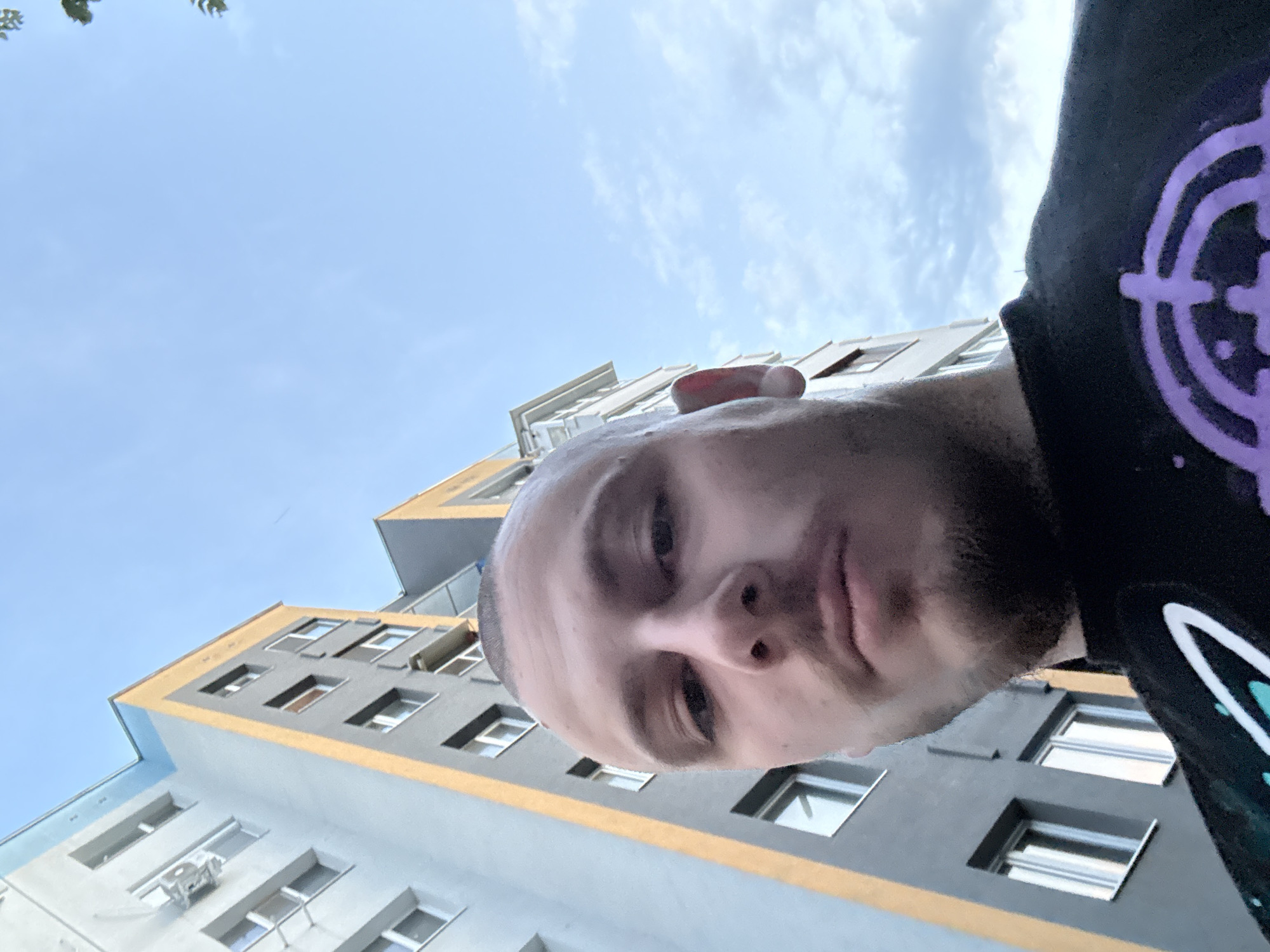 A low-angle self-portrait shows a man in front of a multi-story building, likely taken outdoors. The man has light skin, a shaved head, and a beard. He is wearing a black shirt with a colorful design. Behind him is a building with a gray and yellow facade. The sky is visible in the background, with a mix of blue and clouds. While the city of Stara Zagora, Bulgaria is mentioned, no specific location details are discernible from this image alone to verify that.