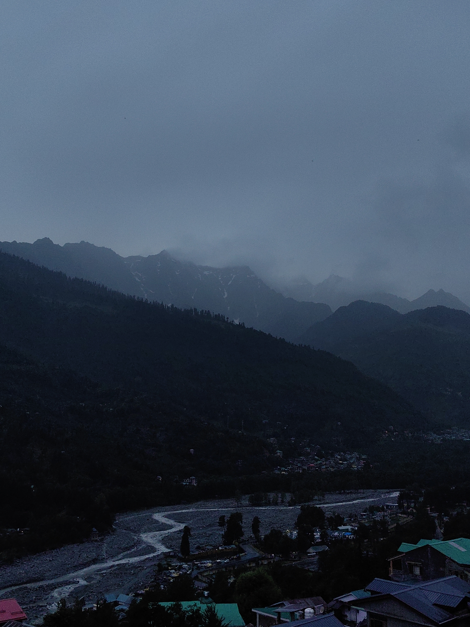 The image depicts a panoramic view of a valley in Vashisht, India, likely during twilight or a heavily overcast day. A winding river, with a rocky bed and shimmering water, flows through the valley floor. On either side of the river, a small town or village is visible, characterized by clusters of buildings with diverse roof colors, including green and blue. Trees are scattered throughout the populated areas and along the riverbanks.
The valley is flanked by steep, densely forested mountains that recede into the distance, fading into a hazy, blue-grey sky. The mountain peaks are silhouetted against the sky, with some showing faint patches of snow. The overall atmosphere is serene and somewhat somber due to the muted color palette and the dramatic mountain landscape. There are no visible people or specific actions taking place. The scene is a natural landscape with human settlement integrated within it.
