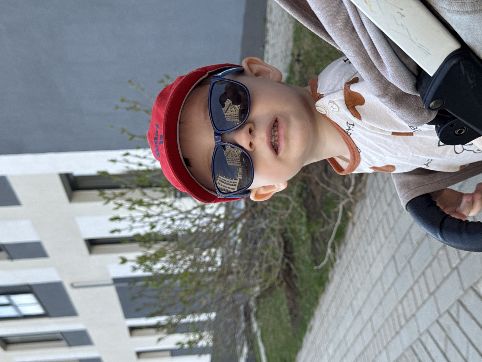 A child wearing a red hat and sunglasses sits in a stroller. The building in the background, reflecting in the child's sunglasses, indicates that the photo was taken in Izhevsk, Russia. The child wears a patterned, light-colored shirt. They are sitting near a young tree on a brick walkway with grass nearby. A building with gray and white panels is visible in the background.