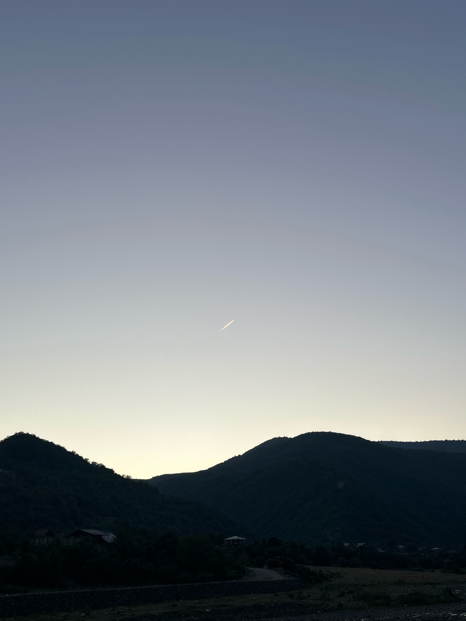 The image depicts a serene natural landscape at dusk in the city of Pavleuri, Georgia. The sky transitions from a deep twilight blue at the top to a pale, warm yellow near the horizon. A faint white contrail from an aircraft is visible against the gradient of the sky. The foreground is dominated by the dark silhouettes of rolling hills covered in dense foliage, suggesting a forest or wooded area. Below the hills, several small, dark shapes indicate scattered buildings, possibly houses, nestled amongst the trees. The lower part of the image shows a dark, uneven ground, possibly a field or rocky terrain, with hints of what might be a dry riverbed or a path. The overall impression is one of tranquility and natural beauty in a rural setting.