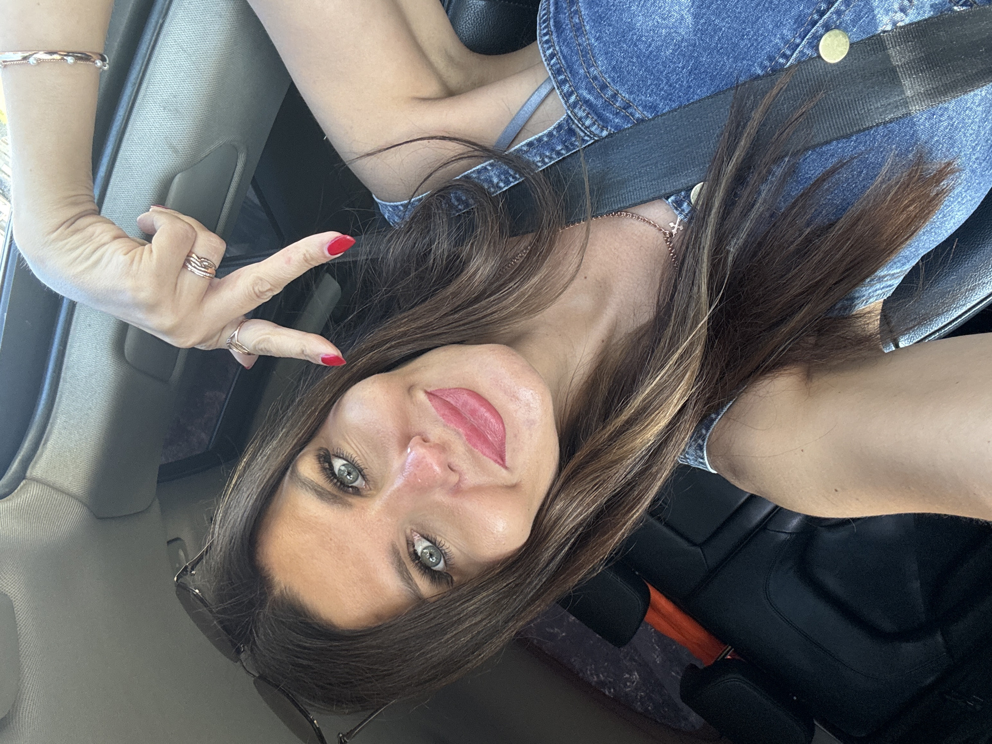 A woman is taking a selfie in a car. She has long brown hair, blue eyes, and is wearing a denim jumpsuit. She is smiling and holding up her hand in a peace sign. The car appears to be in a city environment.