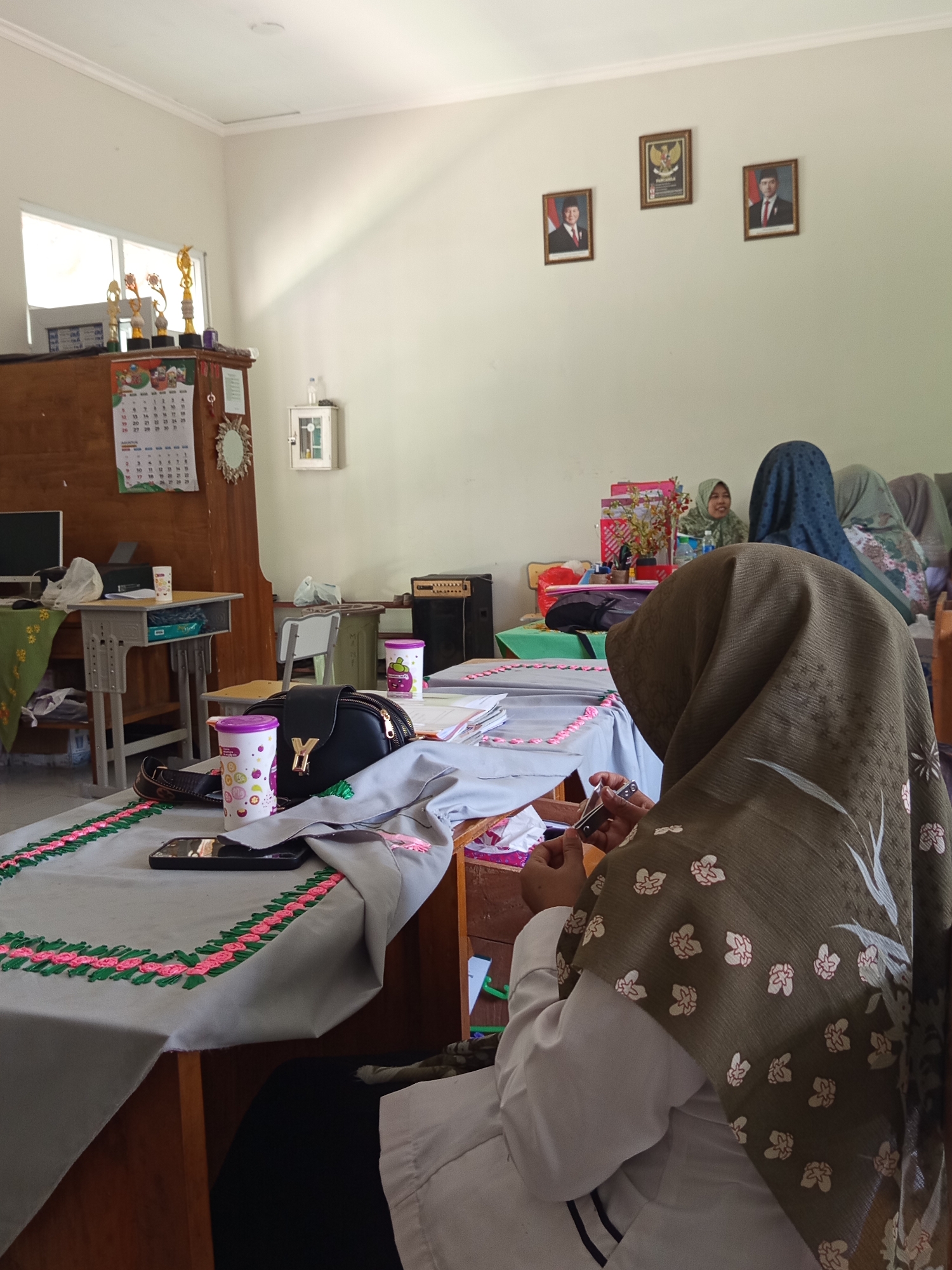 The image depicts an indoor scene, likely a classroom or meeting room, within the city of Lumutan, Indonesia. In the foreground, a woman wearing a brown hijab and a white uniform is seated at a desk, engaged in an activity that appears to be sewing or crafting. She is holding a small silver tool in her right hand and working on a gray fabric adorned with pink and green decorative stitching.
In the background, several other individuals, predominantly women also wearing hijabs, are seated at tables. The room is furnished with desks, chairs, and shelves filled with various items, including trophies, framed pictures of what appear to be Indonesian officials on the wall, and a calendar. A black handbag with a gold "Y" emblem rests on one of the tables, alongside colorful cups and a mobile phone. The environment suggests a structured setting, possibly an educational institution or a community workshop. The lighting is natural, coming from a window on the left side of the image.
