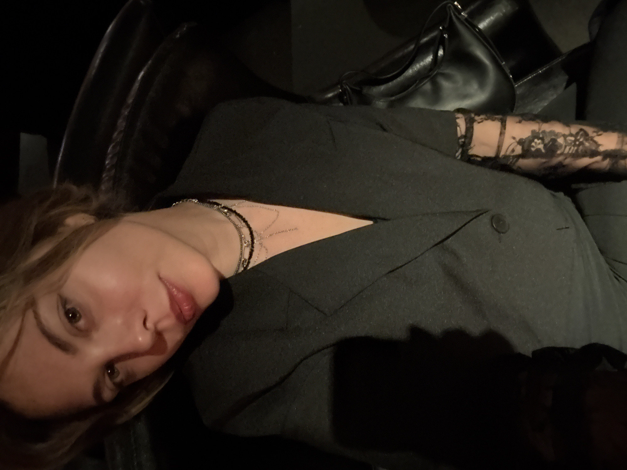 The image shows a close-up, selfie-style photograph of a young woman with light brown hair, looking towards the camera. She is wearing a dark grey blazer, a black top, and multiple layered necklaces. Her right arm is visible, adorned with black lace sleeves. A black leather bag is partially visible behind her. The background is dark and out of focus, suggesting an indoor environment, possibly a dimly lit room or a vehicle. There is no indication of the location being Sinyavino, Russia; the image does not contain any recognizable landmarks or context that would suggest this specific location.