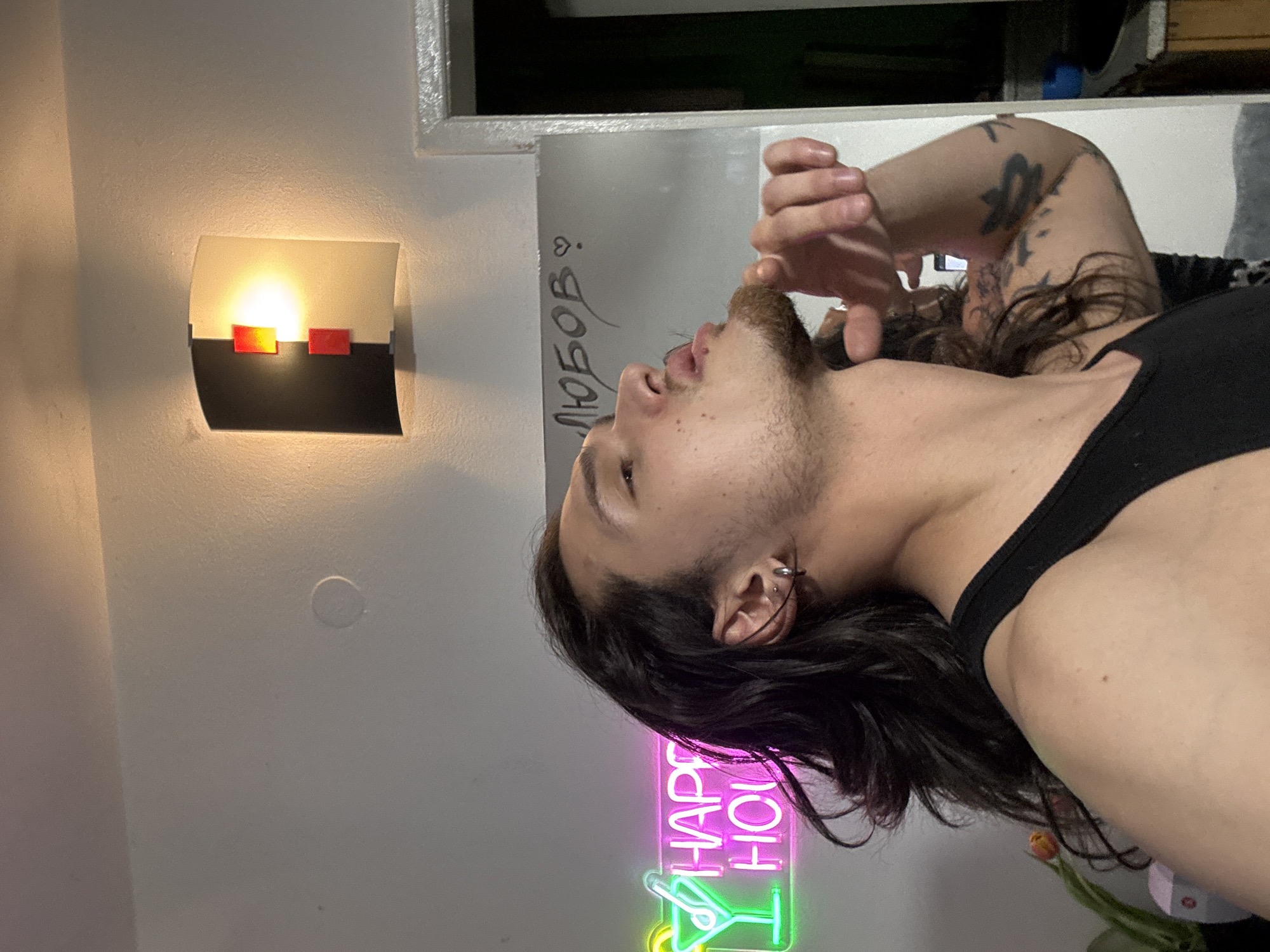 A profile view of a man with long dark hair and a beard, wearing a black tank top, is captured in a low-angle shot. He is looking upward with his mouth slightly open, hand raised.
The background includes: a white wall with a modern sconce on the left; a mirror with Cyrillic writing on it; a "Happy Hour" neon sign with a cocktail glass; and what appears to be a partially visible windowsill or shelf at the top of the frame. It is not possible to ascertain the location in Sofia, Bulgaria.