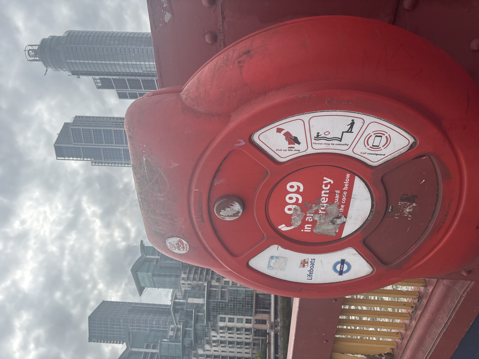 Here is a detailed description of the image:
The image shows a red emergency life ring station, likely situated on a bridge. The station has instructions on how to use the life ring in the event of an emergency, including directions to "Pick up life ring," "Throw ring to casualty," and "Call coastguard." The number "999" is prominently displayed, indicating it's the emergency number to call. Logos for "Lifeboats" and "River" (the London Underground line) are visible on the station. In the background, a view of London skyscrapers through a cloudy sky is present, indicating the location is in London, UK.