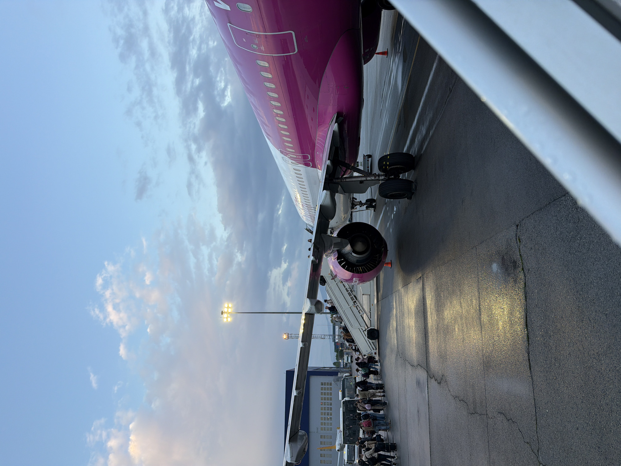 The image depicts an airport tarmac scene, likely during evening or early morning, in Sofia, Bulgaria. A large passenger airplane, identifiable as a Wizz Air aircraft by its distinctive magenta and white livery with "WIZZ" visible on the tail, dominates the frame. The plane is parked on a wet asphalt surface, which reflects the bright artificial lights of the airport, suggesting recent rain or damp conditions.
A prominent activity is the boarding or disembarking of passengers. A mobile boarding stair is positioned at the aircraft's entrance, and a long queue of people extends from it along the tarmac. The individuals in the queue, dressed in a variety of casual clothing, appear to be waiting their turn to ascend the stairs. Some are carrying luggage, such as backpacks.
The sky is partially cloudy, showing a gradient from lighter blue to darker, more overcast tones, consistent with either dawn or dusk. A tall floodlight tower with bright, yellowish lights illuminates the scene from the left, casting long reflections on the wet ground. In the background, beyond the plane's wing and the queue, glimpses of other airport buildings and what appears to be a ground vehicle (possibly a bus) can be seen. The overall setting is a functional, busy airport environment.