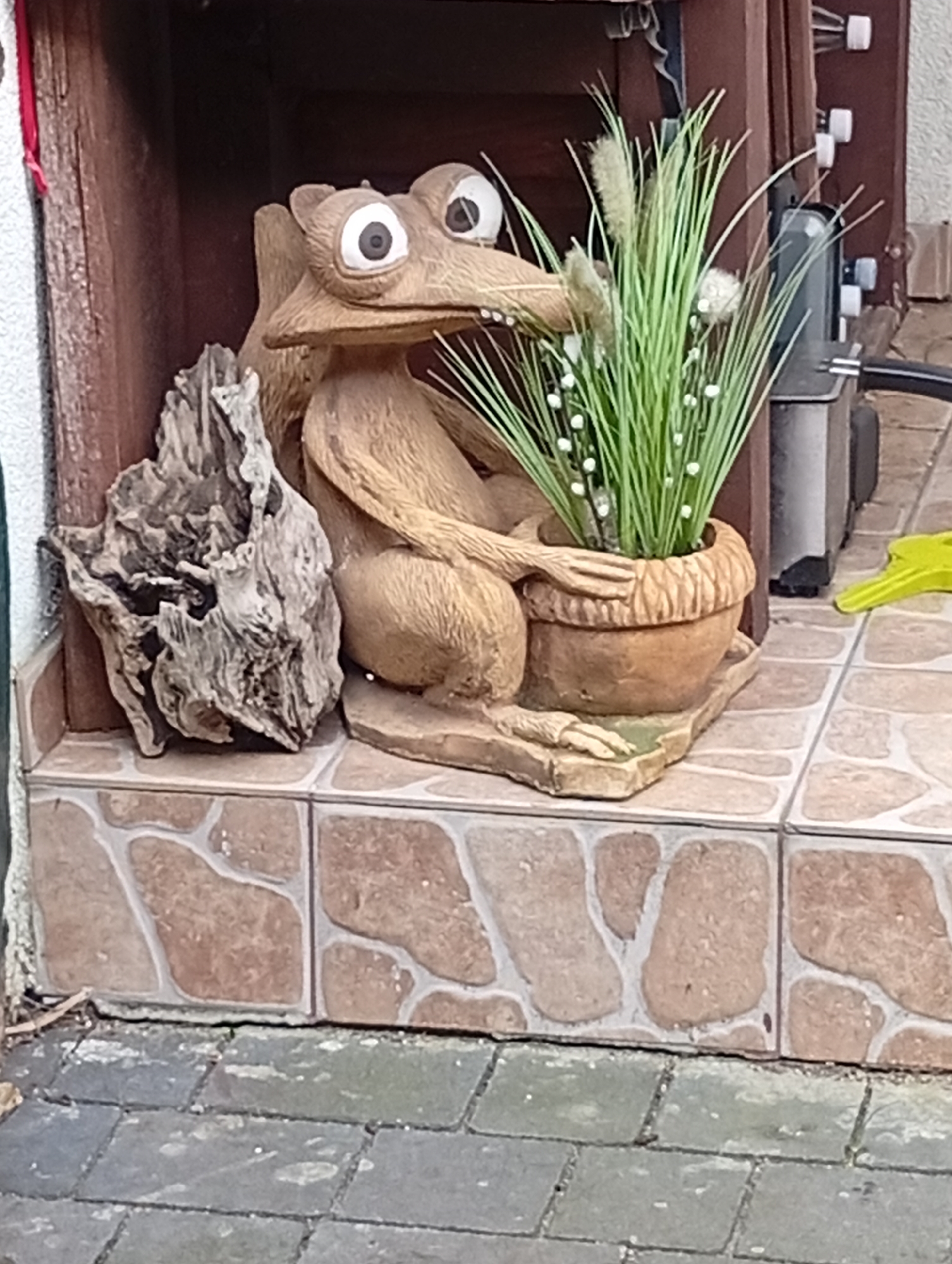 This is a medium-angle, eye-level, full shot of a garden statue situated in front of a building in Kiełczów, Poland. The statue is of a cartoonish, tan-colored creature with large, cartoonish eyes, long ears, and a long snout. It is seated and appears to be holding a terra cotta pot filled with green, artificial grass and small white decorative beads. The statue is positioned on a tiled surface composed of irregularly shaped brown and cream-colored tiles, which are bordered by darker, gray cobblestones. To the left of the statue is a piece of weathered, grey driftwood with a rough texture. The background consists of wooden structures and a metal appliance. The scene is outdoors, likely in a residential setting, given the decorative nature of the statue and the presence of garden elements.