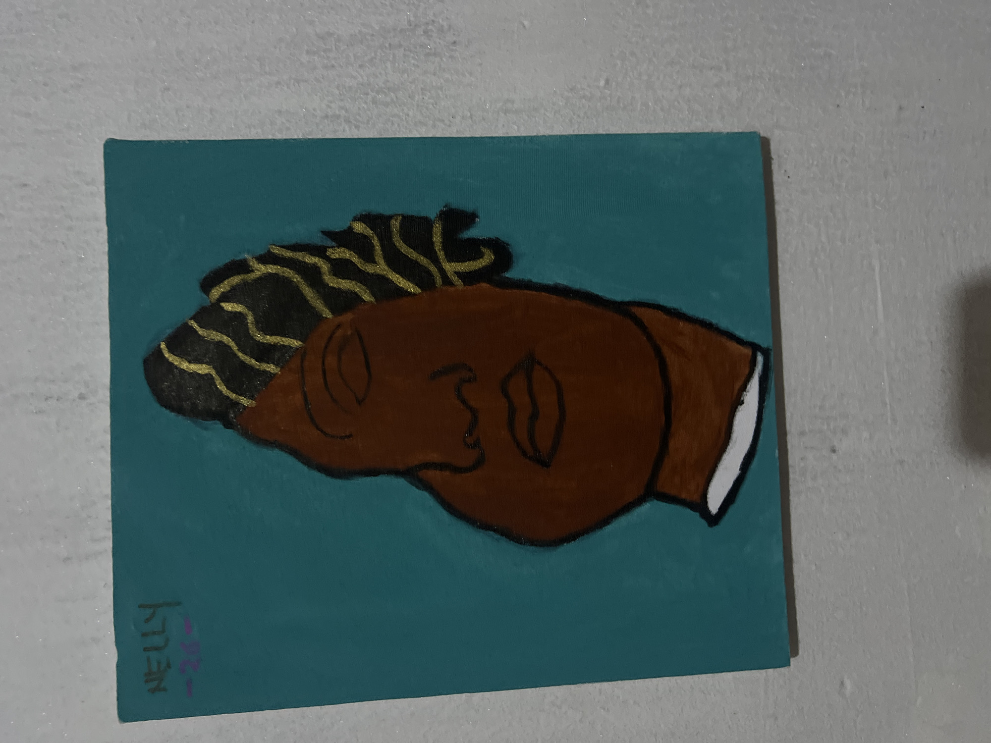 A close-up, slightly tilted, medium shot captures a small painting of a stylized face. The painting is mounted on a turquoise background, and the face is rendered in shades of brown with black and gold accents in the hair. The artwork is signed "NELLY" in the upper left corner, with some unreadable markings below it. The painting is leaning against a textured, light-colored wall.